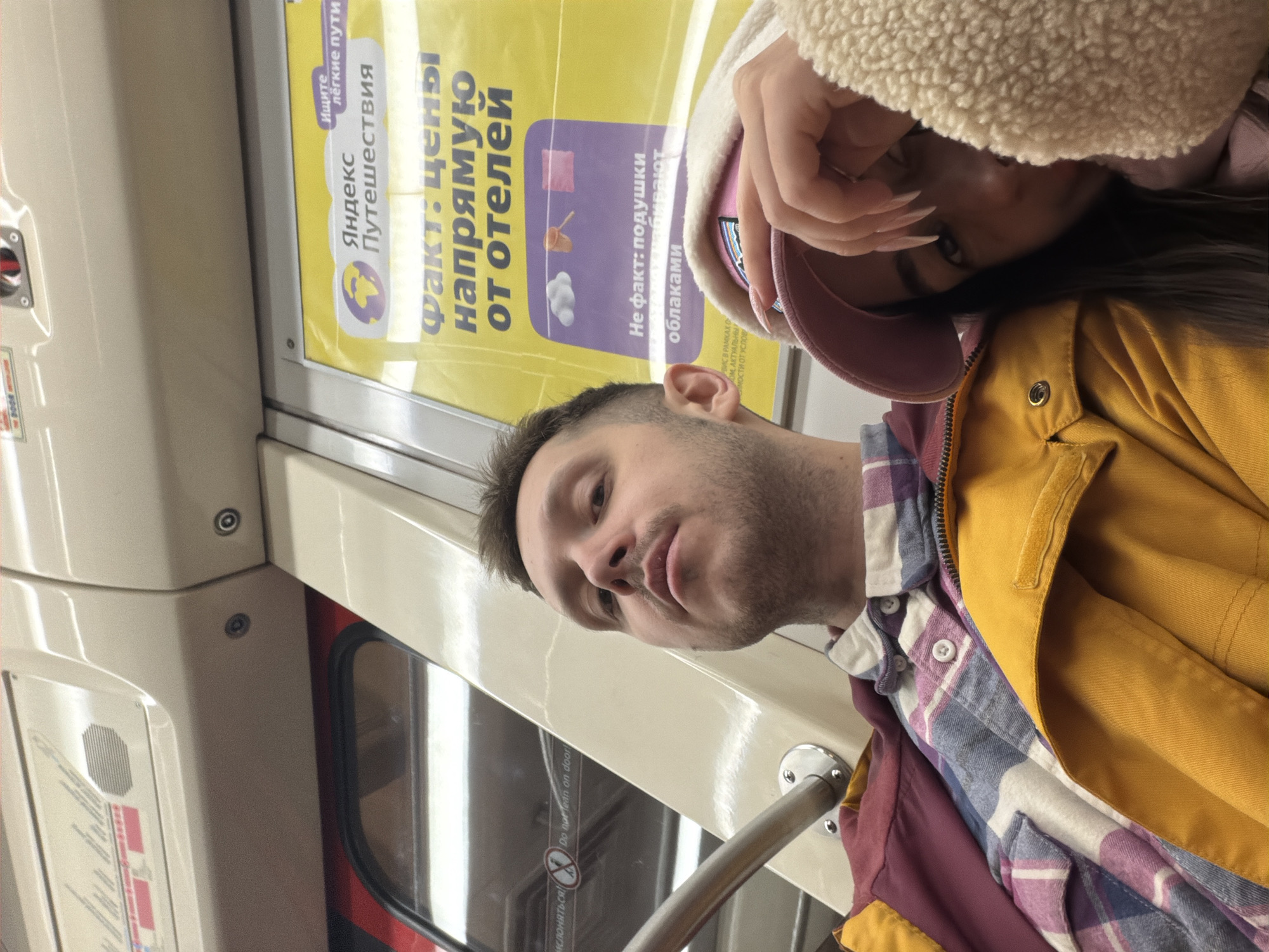 This image appears to be taken inside a subway car. A man is visible, facing the camera, holding onto a handrail. A woman, partially obscured by a fluffy white hood, is behind him. An advertisement is visible on the subway car wall, featuring the logo for "Yandex Travel" and text in Russian. The image is taken at an angle. Without additional information, it is impossible to definitively determine that the image was taken in Saint Petersburg, Russia, although the language of the advertisement suggests a Russian-speaking area.