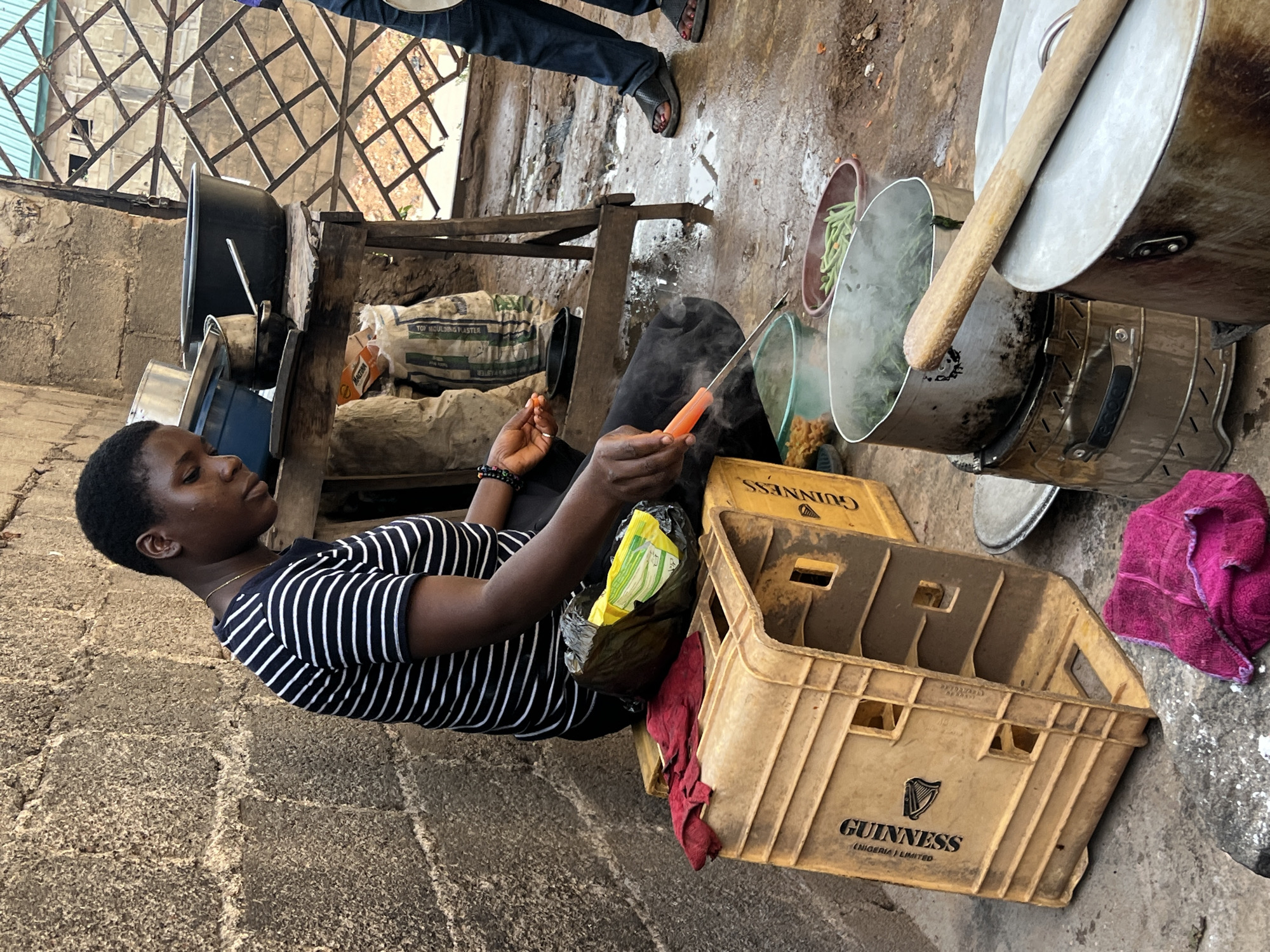 In Jos, Nigeria, a person is sitting outdoors next to a cooking setup. The individual is wearing a striped shirt and dark pants, holding an orange object, possibly a carrot, in their left hand and a knife in their right hand. Next to them is a yellow Guinness-branded plastic crate, with a plastic bag of food on top, and a red rag. Cooking pots are seen with steam coming out of at least one, and a wooden spoon rests on a lid. Other pots and containers are visible, along with what appears to be a bag of plaster. The surroundings are a mixture of stone walls and a metal grille overhead. Another person's legs and feet are visible in the background.