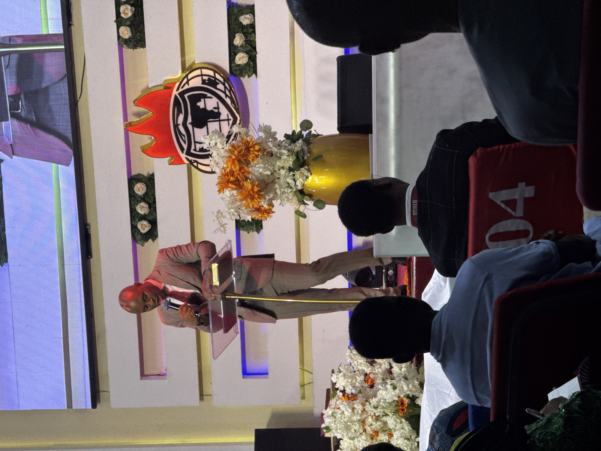An African man in a light brown pinstripe suit and red tie stands at a podium speaking into a microphone. He is bald and has a light-skinned complexion. Behind him, a large screen displays a blurry image, and a logo with flames and a globe is visible on the wall. To the right of the podium, a golden vase holds a bouquet of white and orange flowers. In the foreground, the backs of three people are visible, seated in chairs in what appears to be a large indoor venue. One person in the center wears a light blue shirt and has a lanyard that reads "USHER." The chairs are red with white numbers printed on them, specifically "04" is visible on one. The overall setting suggests a religious or public speaking event. The specific location of Iddo Tudunwada, Nigeria, is not visually identifiable from the image itself.