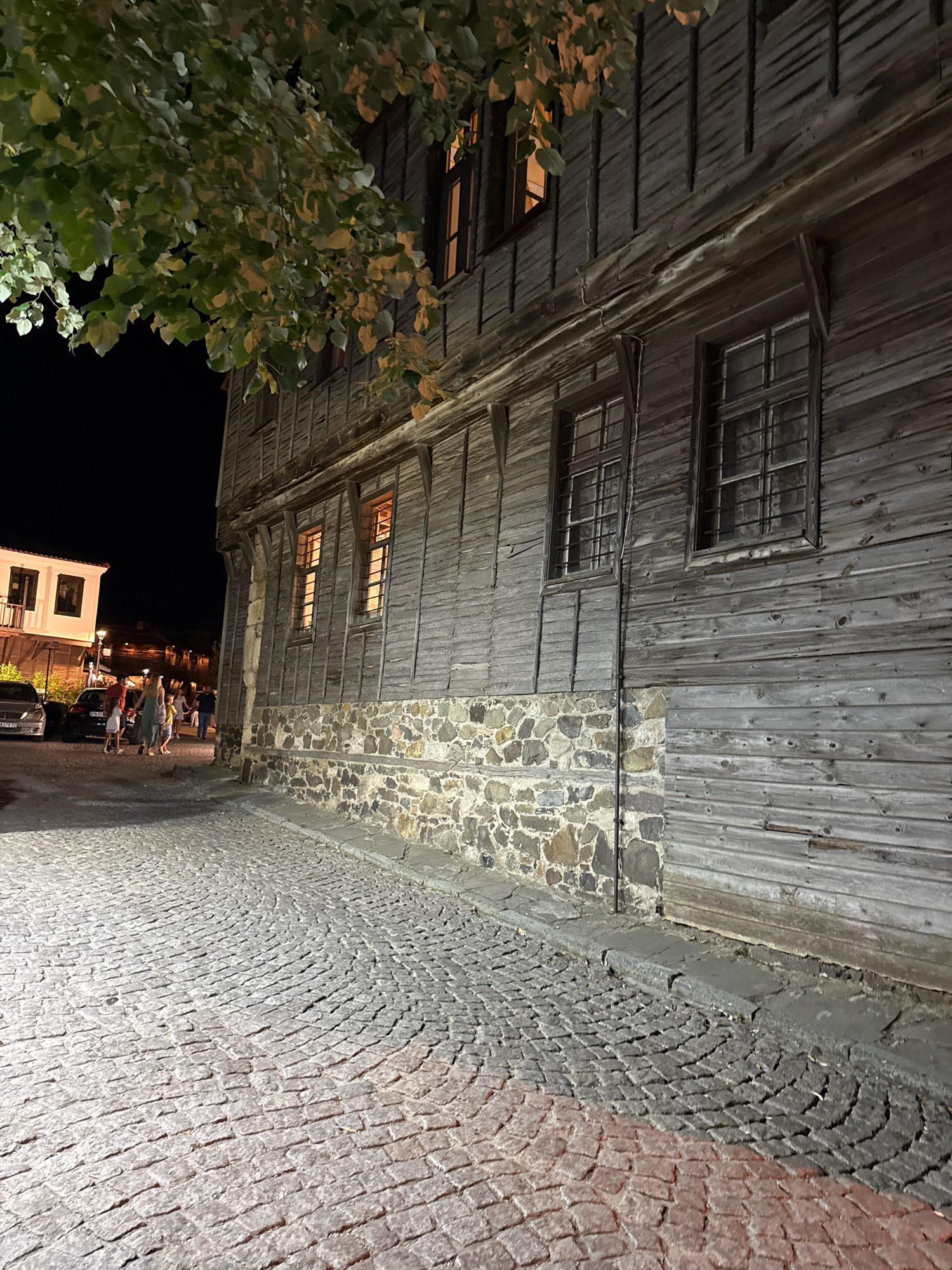 The image depicts a cobblestone street in Sozopol, Bulgaria at night. A large, rustic wooden building with stone foundations dominates the right side of the frame, its weathered planks and barred windows suggesting an older architectural style. Warm light emanates from some of the windows. A leafy tree branch hangs over the top left of the building. On the left side of the street, a smaller, lighter-colored building is visible in the distance, with lights on inside. Several cars are parked along the street, and a few people, including a family with children, are walking. The cobblestones are laid in a curved pattern, with some areas illuminated by artificial light, casting reddish-orange and white glows. The overall atmosphere is one of a charming, historic European town at dusk or night.