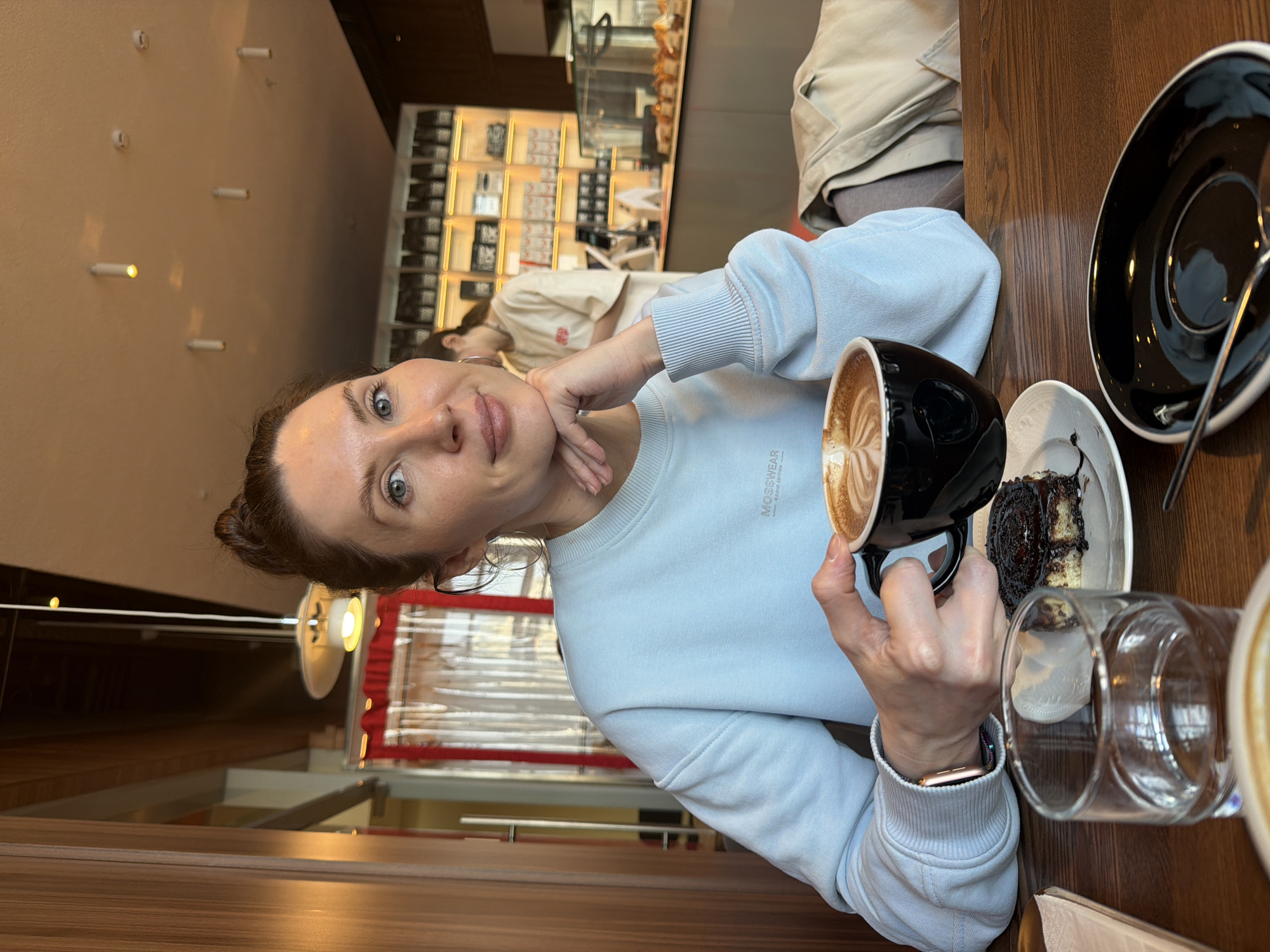 A woman is seated at a wooden table in a cafe. She is wearing a light blue sweatshirt with the word "MOSSWEAR" visible on the upper left. She has light skin, light eyes, and her hair is pulled back. She is holding a black mug with a latte art design in the coffee. On the table, there is a small plate with a piece of chocolate cake, a glass of water, and a black saucer with a spoon. The background shows another person seated, and a display case with items. The cafe appears to be in Perm, Russia.