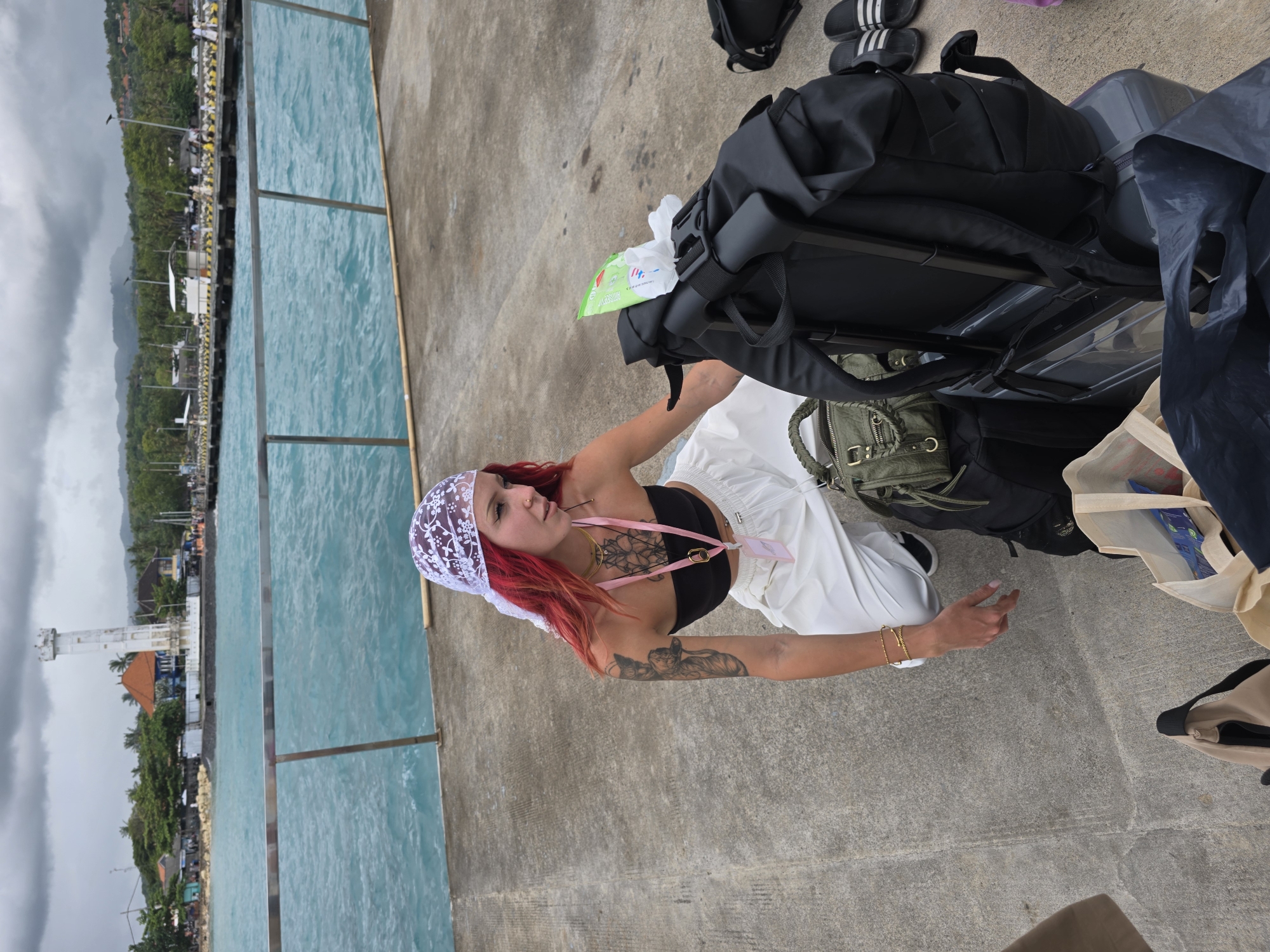 A red-haired woman wearing a white lace headscarf, black top, and white pants, with visible tattoos on her chest and left arm, stands on a gray concrete pier in Jungutbatu, Indonesia. She is surrounded by luggage, including a black suitcase and backpacks. The turquoise water of the sea is visible on the left, separated from the pier by a silver railing. In the background, the coastline is visible, with trees, buildings, and a white tower.