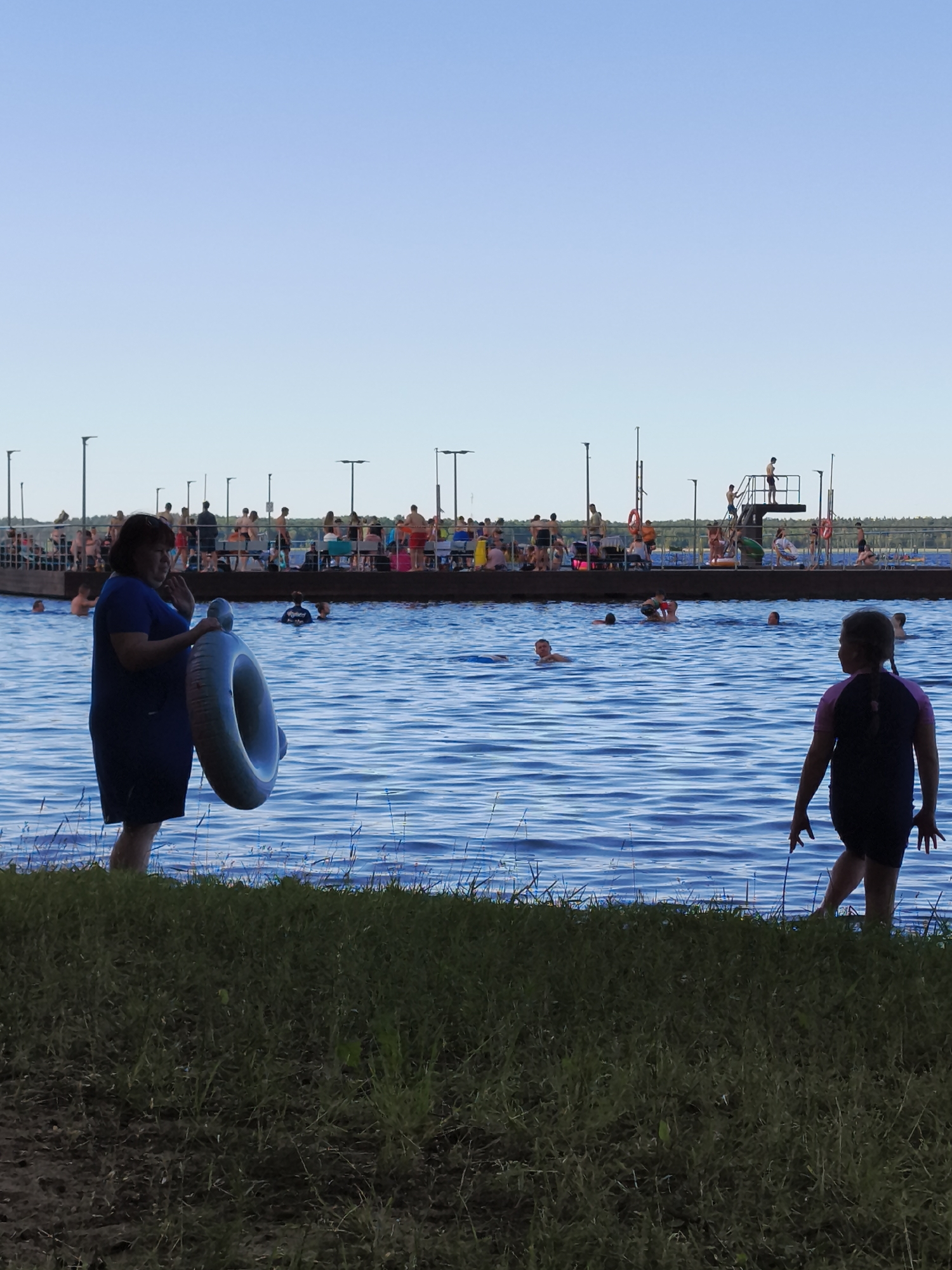 A group of people are enjoying a sunny day at a swimming area. In the foreground, a woman in a blue dress stands by the water's edge, holding a large inflatable ring. To her right, a young girl in a swimsuit is facing away from the camera, looking out at the water.
The water is a vibrant blue, with ripples suggesting gentle waves. Several people are swimming and wading in the water. In the background, a long wooden dock or pier extends into the water, lined with people sitting and standing. A diving platform with a ladder is also visible on the dock, with individuals preparing to jump or having just jumped. A few people are visible on the dock or in the water near it.
The sky is a clear, bright blue, indicating good weather and likely daytime, possibly late afternoon given the lighting. Lampposts are visible along the edge of the dock and in the distance, suggesting this is a developed recreational area. The overall scene conveys a sense of leisure and summer fun. No visible text is present in the image.