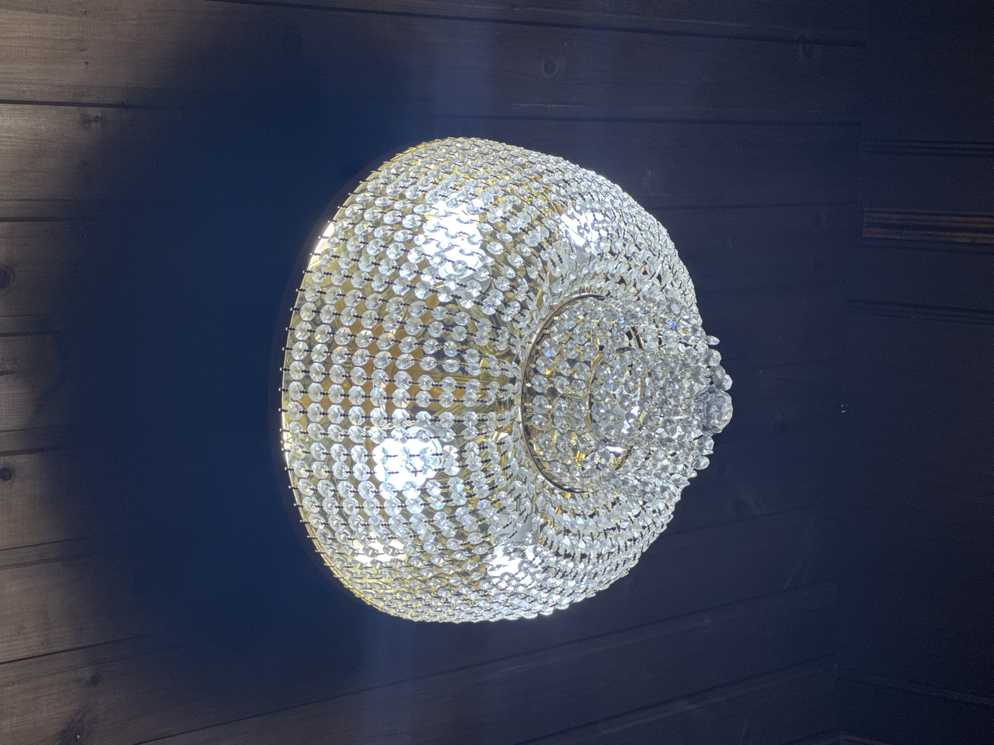 A crystal chandelier hangs from a dark wooden ceiling. The chandelier has a circular, multi-tiered design with numerous faceted crystals reflecting light from internal bulbs. The ceiling is constructed of parallel wooden planks, running diagonally from top left to bottom right. The background is a deep, dark blue. There are no people or animals visible in the image. The location is the city of Luch SNT (Putilovo), Russia.