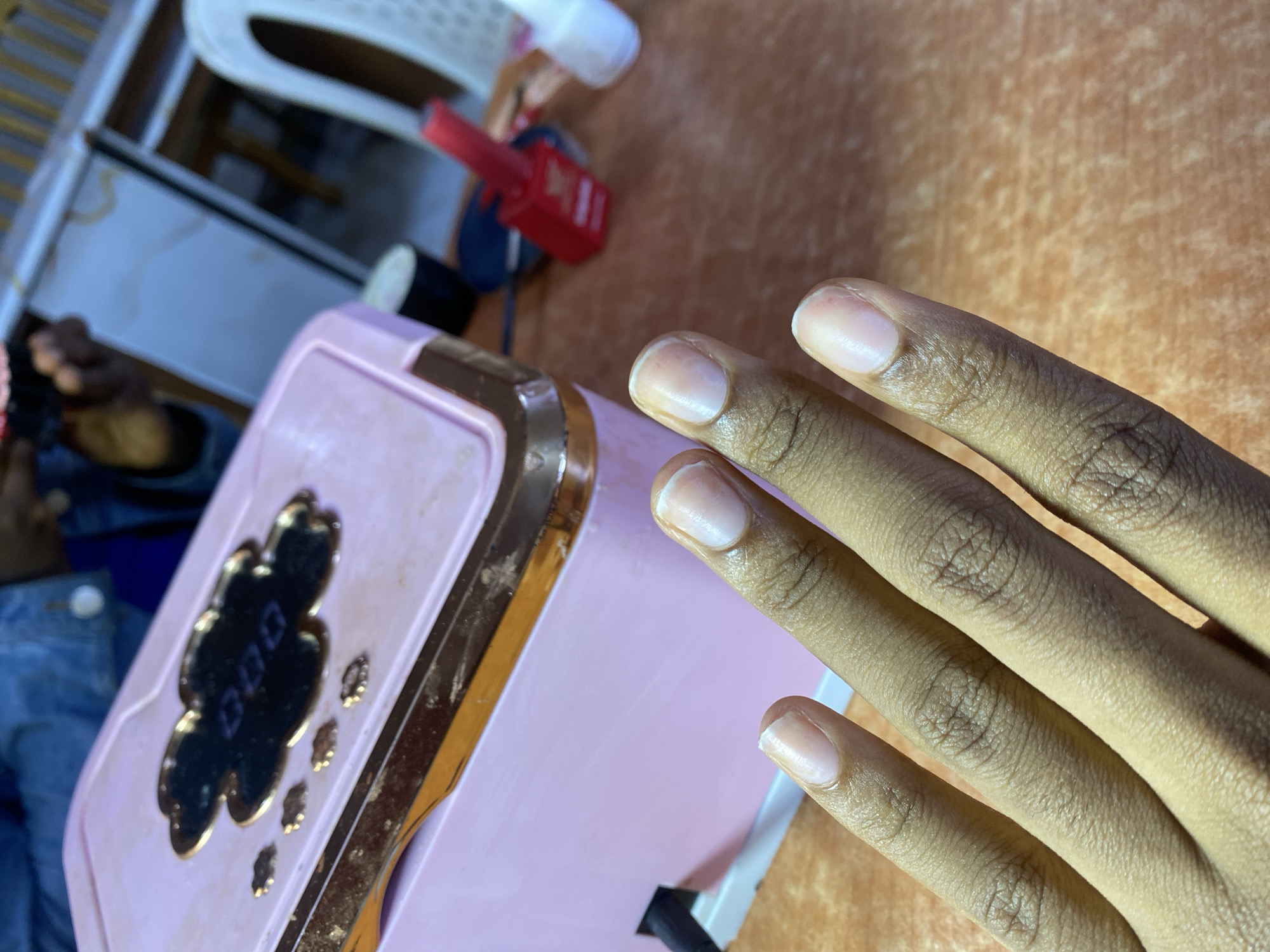 The image depicts a close-up of a person's hand with unpolished nails, positioned in front of a pink nail dryer. The nail dryer has a cloud-shaped design with a gold trim and a digital display showing "000". In the background, a red nail polish bottle is visible, along with other unidentifiable objects and furniture, suggesting an indoor setting, likely a nail salon. The background also shows a hint of what appears to be a white chair. The lighting suggests an indoor environment, possibly during the daytime. There is no explicit indication of the city of Jos, Nigeria, within the image itself.