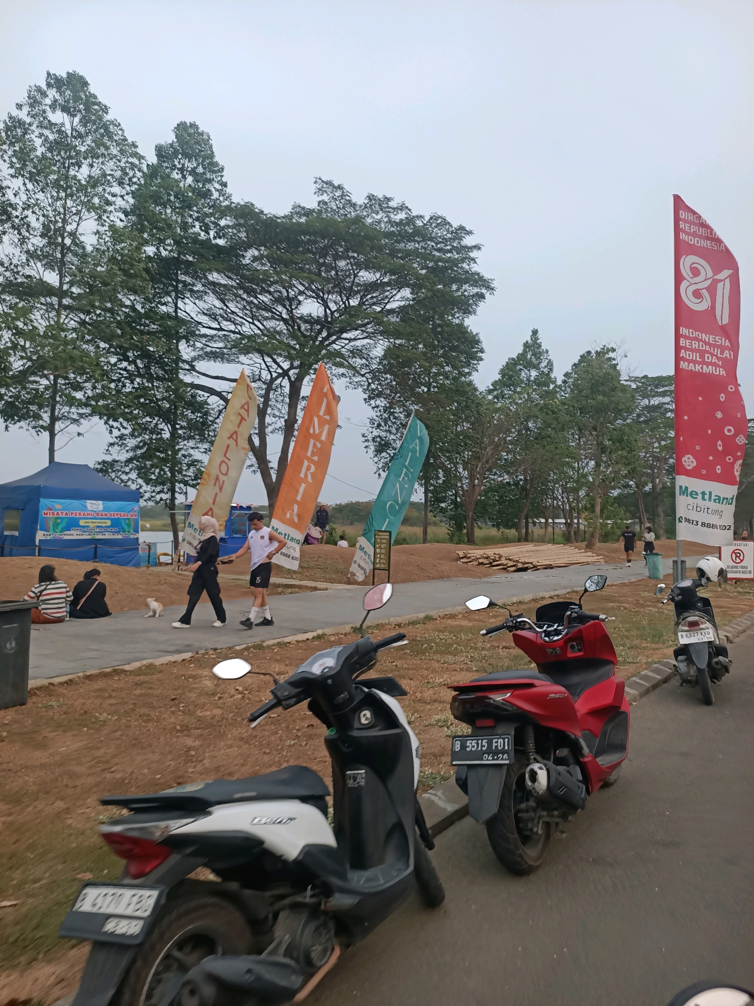 In Telagamurni, Indonesia, on a cloudy day, a scene unfolds in an outdoor recreational area. In the foreground, two scooters are parked on a paved road. The scooter on the left is white and black, with its rear visible. The scooter on the right is red and black, with a license plate reading "B 5515 F01". Behind the scooters, a pathway borders a grassy area with scattered trees.
Further back, several colorful banners or flags stand tall, displaying text that is partially legible. To the left, a blue tent structure with signage is set up, possibly for information or an event. In front of the tent, a small white dog walks towards a young couple. The couple, a woman in black Islamic attire and a man in athletic wear, are walking away from the viewer. Other individuals are scattered throughout the midground, some sitting and others walking. In the background, lush green trees dominate the landscape, with a hint of a body of water visible beyond them. To the far right, a tall red banner displays "DIRGAHAYU REPUBLIK INDONESIA 81" and other text.