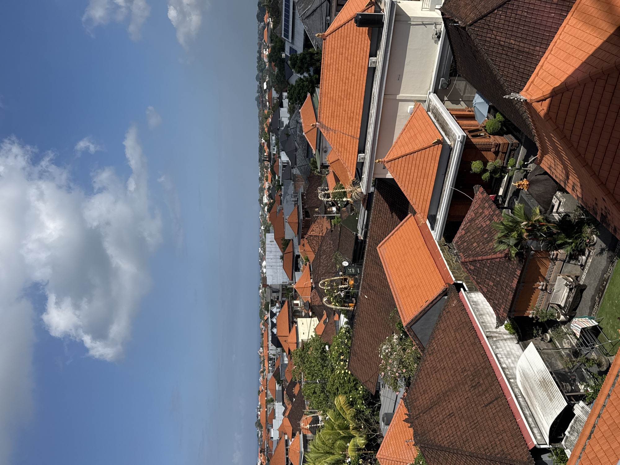 This is a high-angle, full-shot photograph of a dense residential area in North Kuta, Indonesia, under a bright blue sky with scattered white clouds. The scene is dominated by numerous rooftops, primarily covered with terracotta tiles in varying shades of orange and brown. The architecture suggests a tropical, possibly Balinese, style with distinct pitched roofs, some with decorative finials.
In the foreground, several prominent rooftops are visible, including a large one covered in darker brown tiles and another with vibrant orange tiles. A white, corrugated metal roof is also present in the lower left quadrant. Beyond these, a sprawling cityscape unfolds, characterized by a continuous expanse of similarly tiled roofs, interspersed with greenery from trees and palm fronds. Some buildings feature white or light-colored walls.
In the mid-ground, specific structures are discernible, including one with a prominent orange-tiled roof and white walls, featuring several windows and what appears to be an air conditioning unit. There are also smaller, intricately designed buildings with dark wooden elements and decorative roof structures. Some of these buildings have outdoor seating areas with benches and small trees or potted plants. In one courtyard, a light blue umbrella is visible.
Further in the distance, the cityscape stretches towards the horizon, where a faint line of buildings and possibly the ocean can be seen. Solar panels are visible on some of the rooftops in the mid-to-far distance, indicating a mix of traditional and modern elements. The overall impression is one of a well-established, densely populated urban or semi-urban environment. No people are visible in the frame. The lighting suggests it is daytime, with the sun casting shadows on the rooftops, indicating the sun is at an angle.