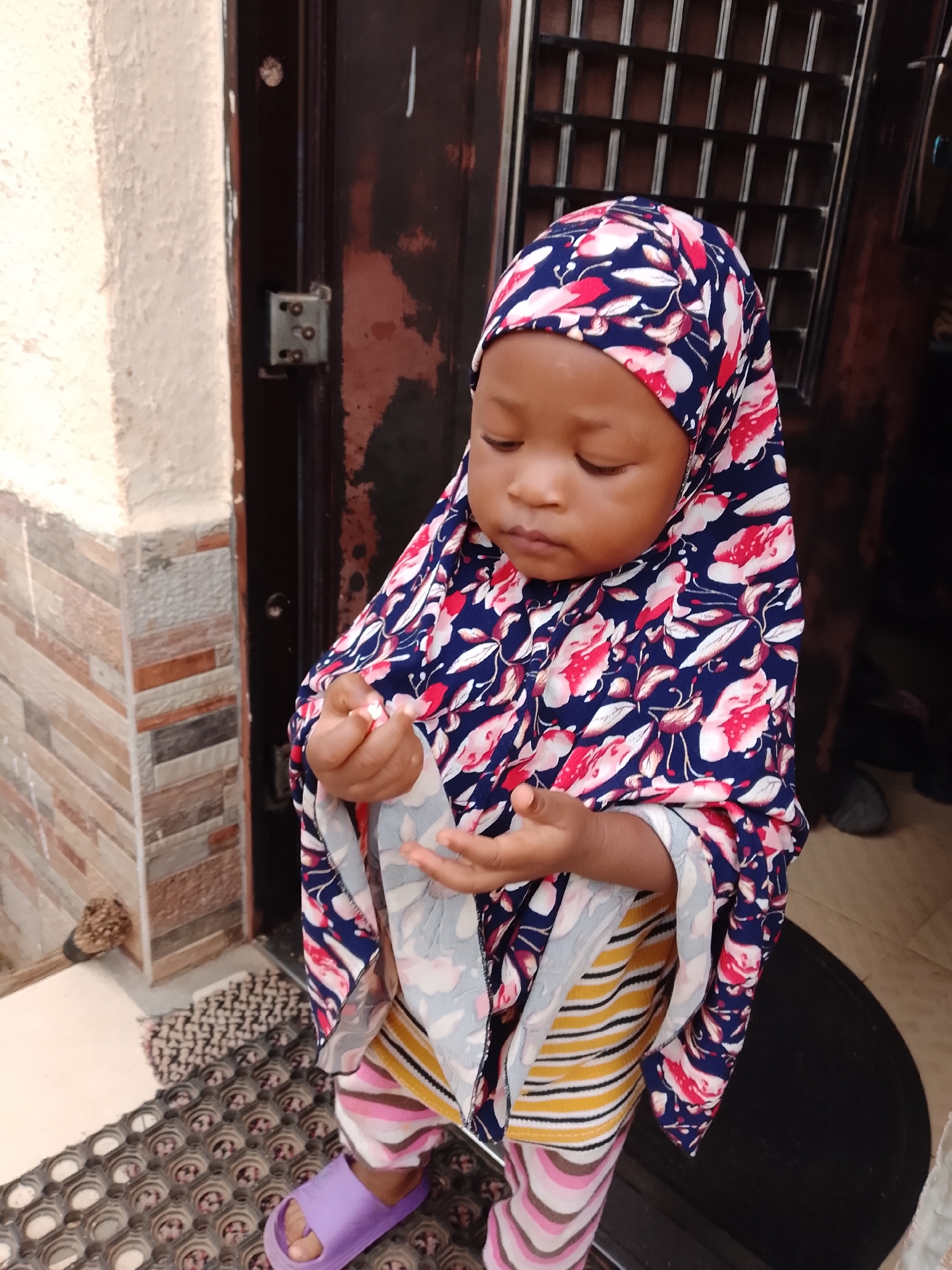 A young child, likely a girl, stands near a doorway in Jos, Nigeria. She is wearing a navy blue hijab with a floral pattern of red and white flowers and leaves, and a white underscarf. Her clothing includes a striped top with yellow, white, pink, and brown horizontal stripes, and pink and white zigzag patterned leggings. She wears a pair of light purple slide sandals.
The child is looking down at her hands, which are clasped together, suggesting she might be playing with something small or examining her fingers. She stands on a patterned mat, possibly a doormat, with an egg carton-like texture beneath it.
Behind her is a dark brown door with a metal grate. To the left of the door is a wall with a light beige stucco finish above a section of brickwork featuring horizontal bands of various earthy tones, including beige, brown, and grey. A metal door lock mechanism is visible on the door frame.
The overall environment suggests an outdoor residential setting in a city.