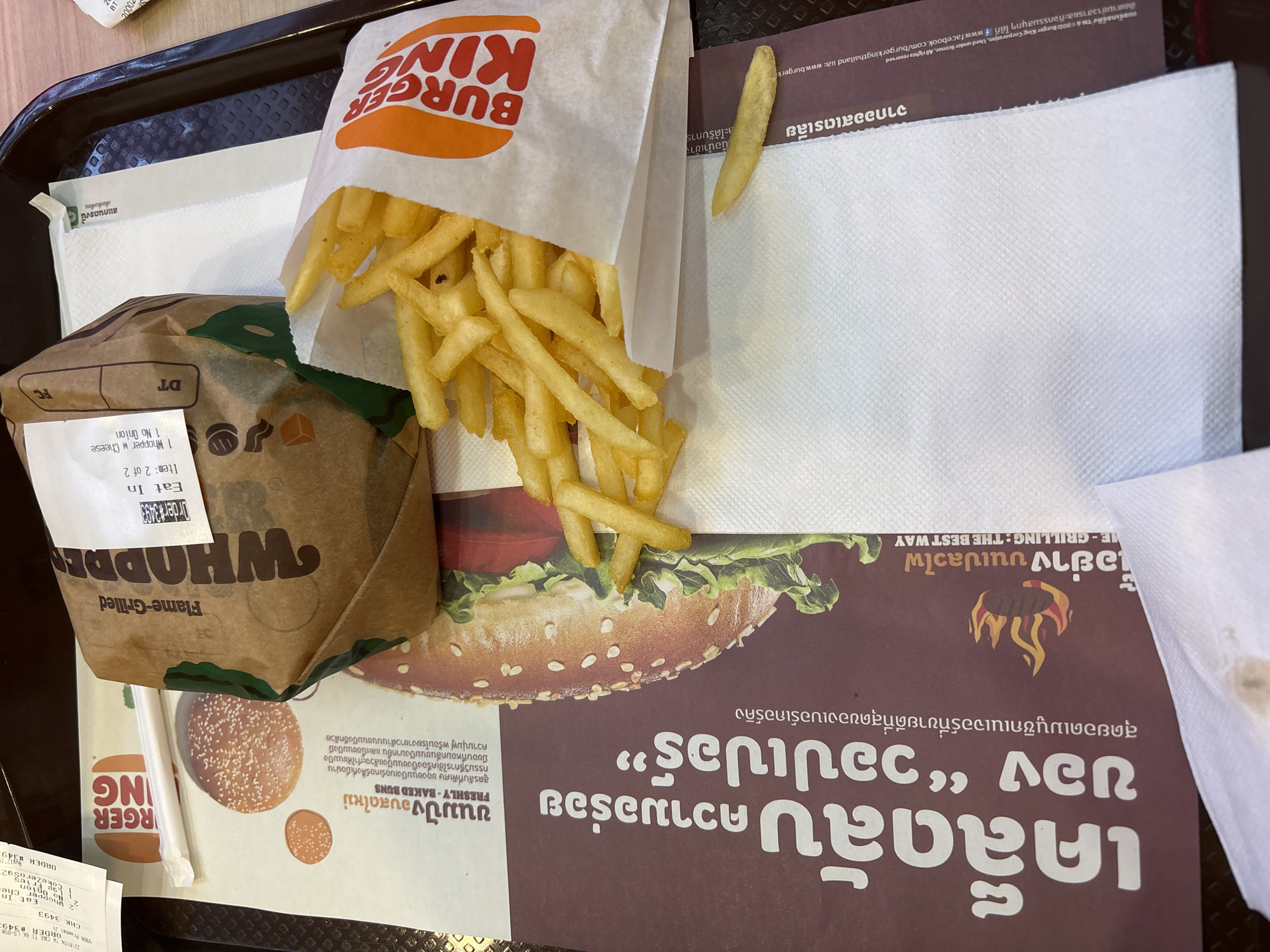 A close-up, overhead shot shows a Burger King meal on a dark gray tray. The meal includes a Whopper sandwich in a brown paper wrapper, a white paper bag filled with french fries, and a white paper napkin. The Burger King logo is visible on the wrapper and the french fry bag. The background is a brown and white patterned placemat with Thai text. The scene appears to be in a Burger King restaurant, likely in Suthep Town Municipality, Thailand, given the Thai text and the presence of Burger King. No people are visible in the image.