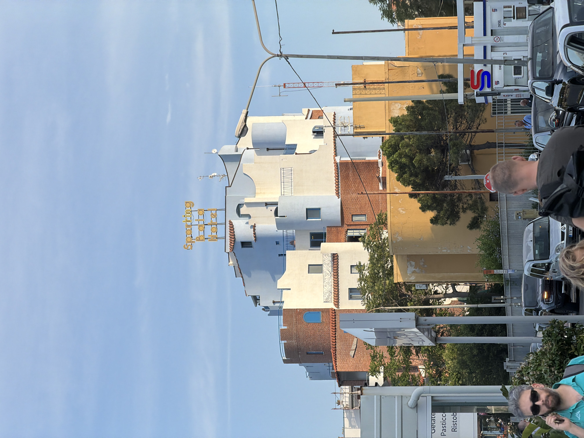 The image depicts a street scene in Chianchitta, Italy. The sky is light blue with faint clouds. On the left side of the image is a multi-story building with a varied facade: sections are pale blue and light beige, while others are brick red. The building has a sign that reads "Sporting Club Hotel". To the right of this building are yellow, weathered buildings. Several trees are interspersed between the buildings. Cars are parked along the street. Several people are visible including a man with a backpack, a man in a green shirt with sunglasses, and other people blurred in the background. Various street signs and shop signs are also visible.