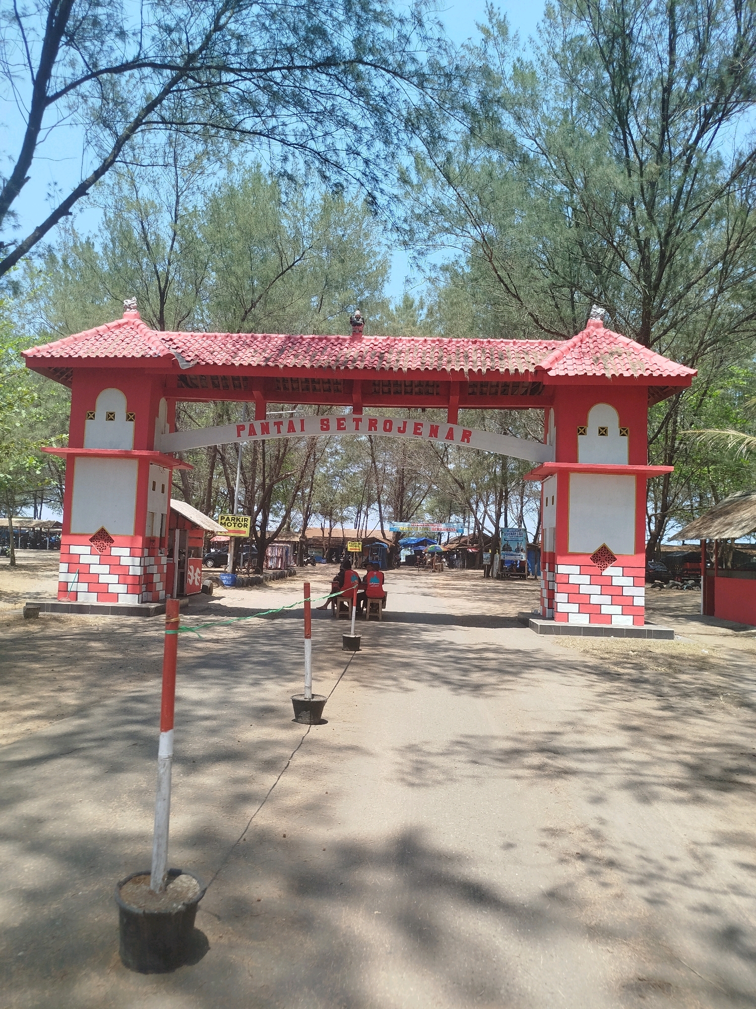 This image captures the entrance to Pantai Setrojenar, a beach in Setrojenar, Indonesia. A vibrant red and white archway, adorned with a red tiled roof and decorative elements, stands prominently in the foreground. The archway bears the inscription "PANTAI SETROJENAR" in white lettering.
The ground leading up to the arch is paved with asphalt, with red and white striped poles on the side, some in black pots. Beyond the archway, the scene opens up to a dusty, sandy area dotted with trees, some of which have sparse, needle-like foliage. In the background, there are several small structures, possibly vendor stalls or small buildings, with thatched roofs and some tarpaulin coverings. A few people are visible, one sitting on a bench near the archway, and others further back among the stalls. A red vehicle and what appears to be a parked motorcycle are also present. The sky above is clear and blue, suggesting a sunny day. The overall impression is that of an entrance to a coastal recreational area.
