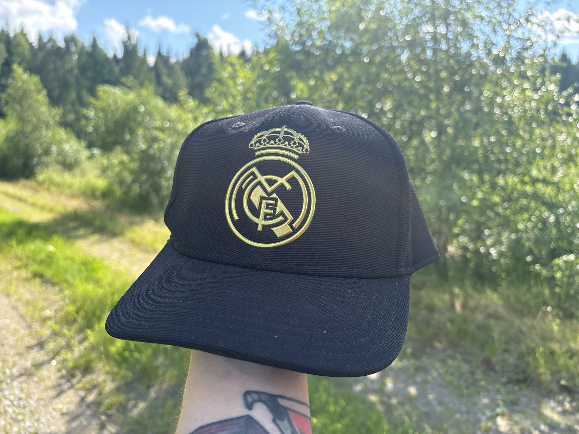 A person's arm is holding up a black baseball cap featuring the gold Real Madrid C.F. crest, which includes a crown and the letters "MCF." The background is a softly blurred, sunlit scene of greenery, likely trees and grass, with a clear blue sky with white clouds above. The sun appears to be high in the sky, suggesting it is daytime, and the lighting indicates fair weather. A dirt path or road is visible to the left of the cap. The person's arm has a tattoo. The overall impression is one of being outdoors in a natural, possibly wooded, setting.