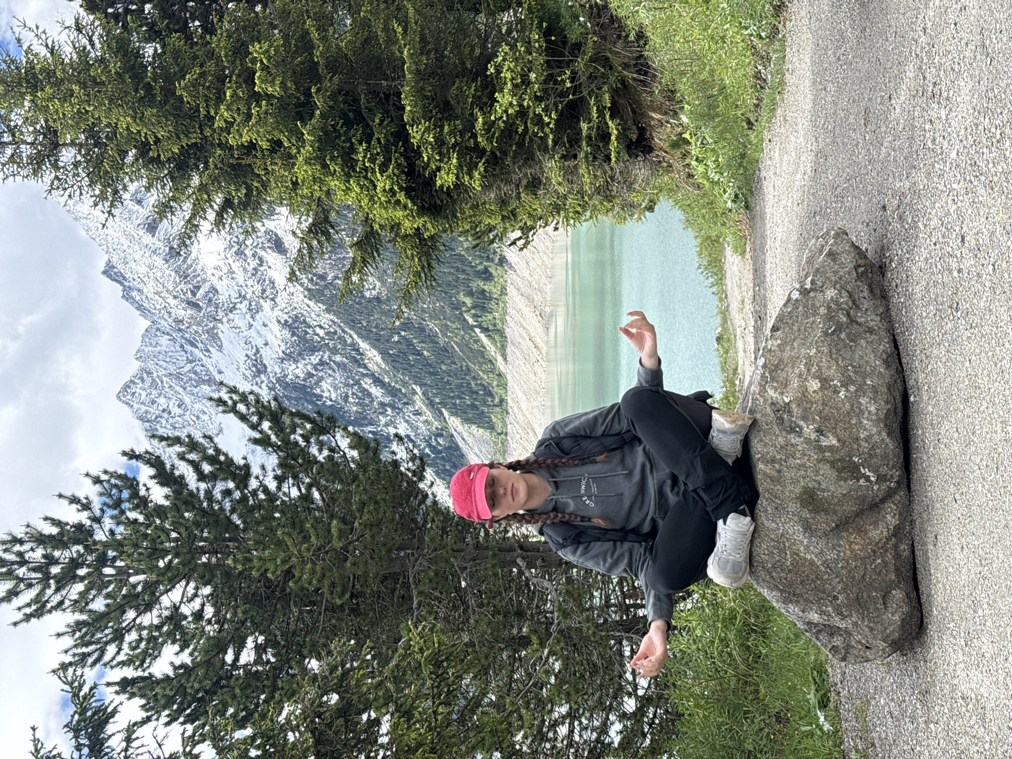 A young woman with braided hair, wearing a red cap, grey hoodie, and black pants, sits cross-legged on a large rock in a meditative pose. She is outdoors with a serene expression and her eyes closed. Behind her, a body of water with a turquoise hue is visible, framed by lush green trees on either side. In the distance, majestic snow-capped mountains rise against a cloudy sky. The ground is a mix of gravel and grass, suggesting a natural setting, possibly a path or trail. The image appears to be taken in Finkenberg, Austria, given the characteristic Alpine landscape.