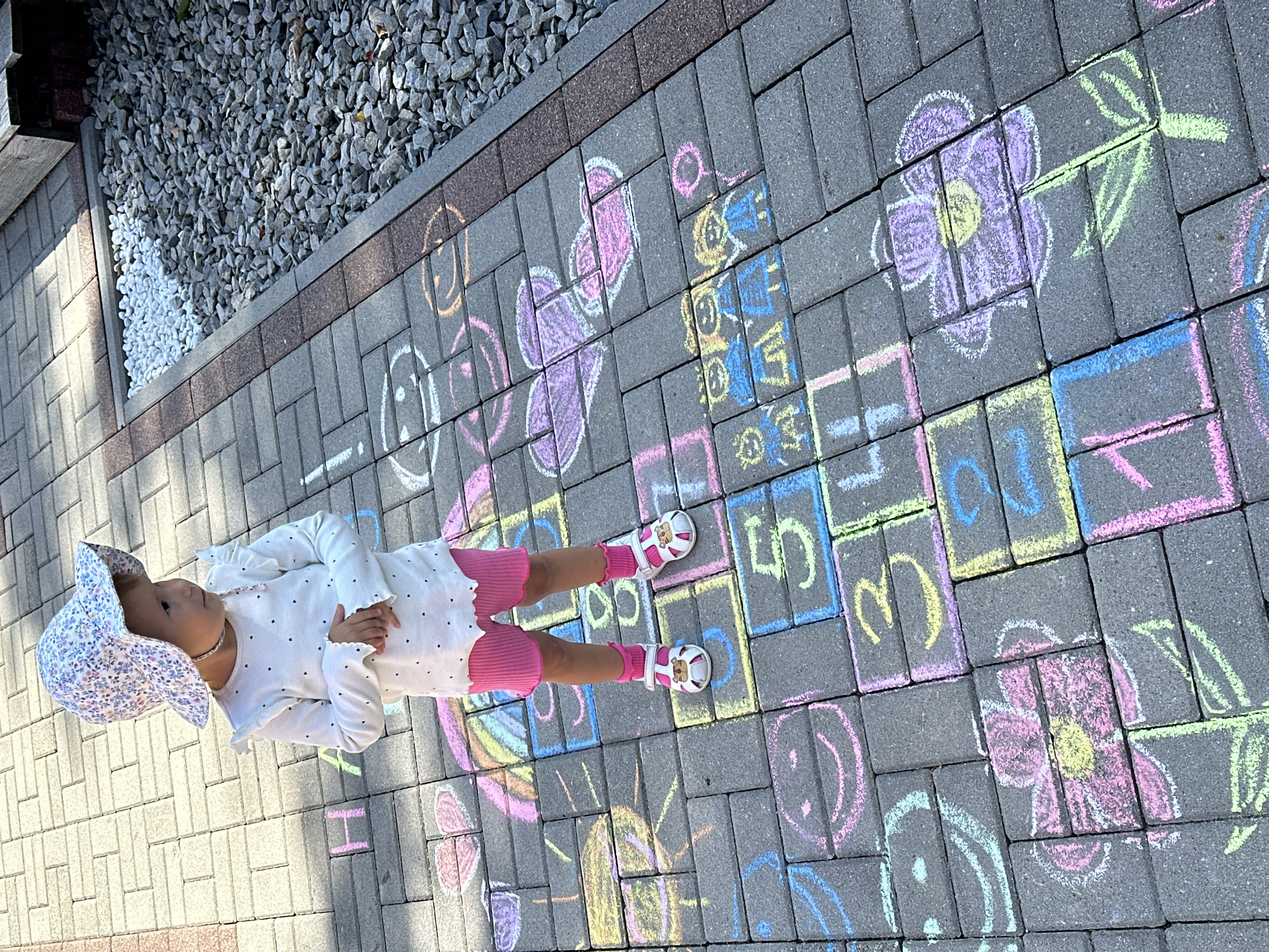 The image depicts a young child standing on a paved area decorated with chalk drawings. The child is wearing a floral sun hat, a long-sleeved white shirt with small black polka dots, pink shorts, pink socks, and white sandals with a tiger design. The paved area appears to be a pathway or patio, made of rectangular grey pavers. Chalk drawings on the pavers include hopscotch numbers (1 through 5 are clearly visible), flowers, hearts, and smiley faces. The context suggests this is an outdoor recreational space. The scene is set in Wisła, Poland, likely a backyard or public park given the hopscotch drawing.