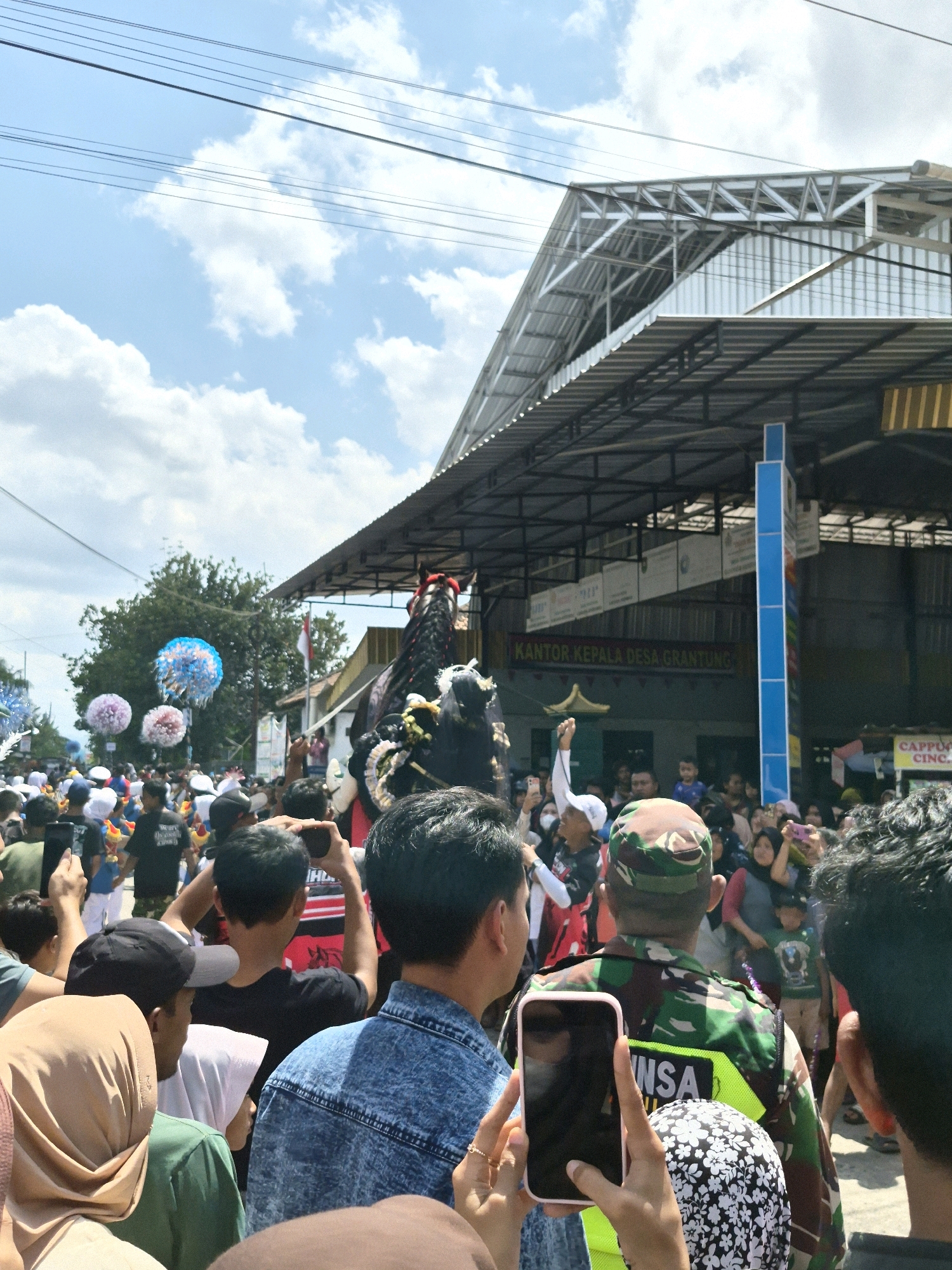 The image captures a daytime outdoor street scene in Grantung, Indonesia, possibly during a parade or festival. A large crowd of people is gathered, many of whom are holding up mobile phones to take pictures or videos. In the center of the frame, a large, ornate costume, possibly depicting a horse or mythical creature, is visible. The costume is black and adorned with decorative elements, including what appears to be a red headdress or mane. Behind the crowd and costume, a building with a corrugated metal roof and a prominent blue sign displaying text in Indonesian is present. The sign reads "KANTOR KEPALA DESA GRANTUNG," indicating this is the Grantung Village Head's office. Several trees are visible in the background, and the sky is blue with scattered white clouds. The overall atmosphere appears festive and crowded.