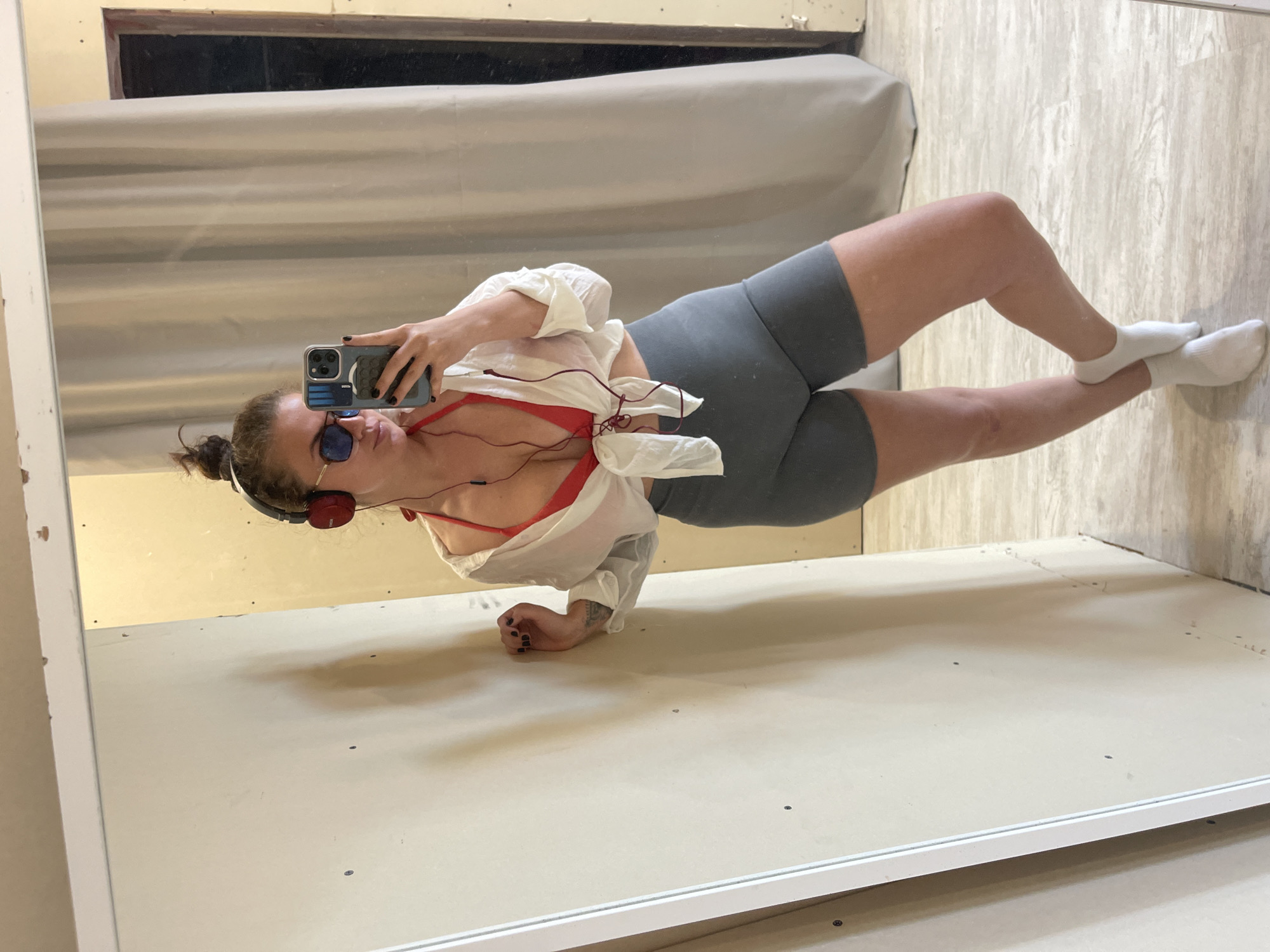 A woman is visible in a mirror taking a selfie, wearing sunglasses, headphones, a red top under a white, unbuttoned shirt, grey shorts, and white socks. She is lying on her left side, propped up on her left arm, with her right leg raised. The background includes a light-colored wall and a wooden floor. The mirror's frame is partially visible. There is no evidence to support the stated location.