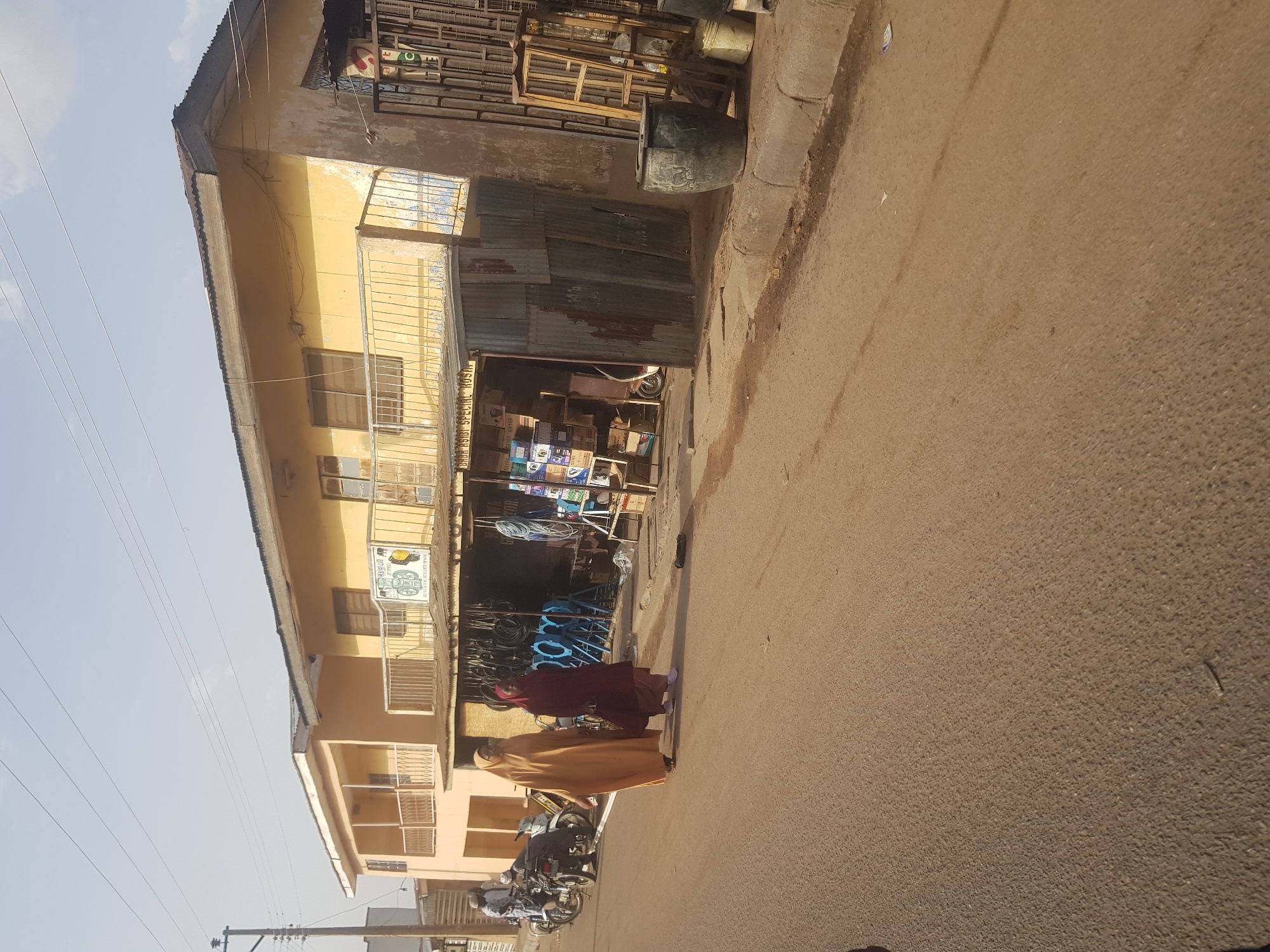 Two women in long dresses walk on the street in Dutsinma A, Nigeria. One woman is wearing a maroon dress, and the other is wearing a tan dress. A motorcycle is parked on the side of the street. Behind them is a two-story yellow building with a storefront, where goods are displayed. The sky is bright and clear.