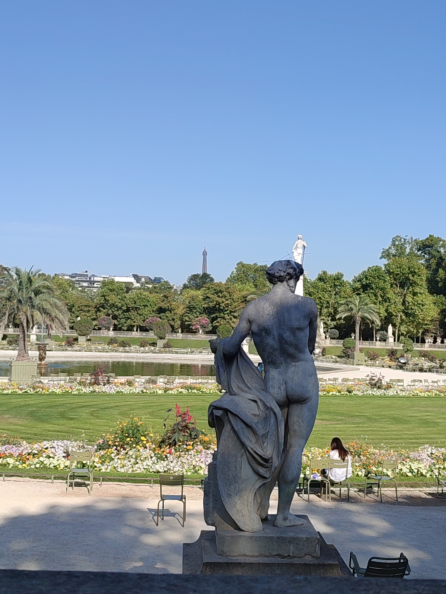 The image depicts a rear view of a statue of a nude figure in a garden with a pond and trees in the background. The Eiffel Tower is visible in the distance, indicating the location is Paris, France. In the middle ground, there is a pond surrounded by a manicured lawn and flower beds. Several green chairs are placed around the lawn, with one woman sitting on a chair in the right middle ground, facing away from the camera. The statue itself is dark-colored and appears to be made of bronze or a similar material, with drapery covering part of the figure's lower body. The sky is clear and blue. The foreground shows a gravel path and the base of another structure, likely a pedestal for the statue.