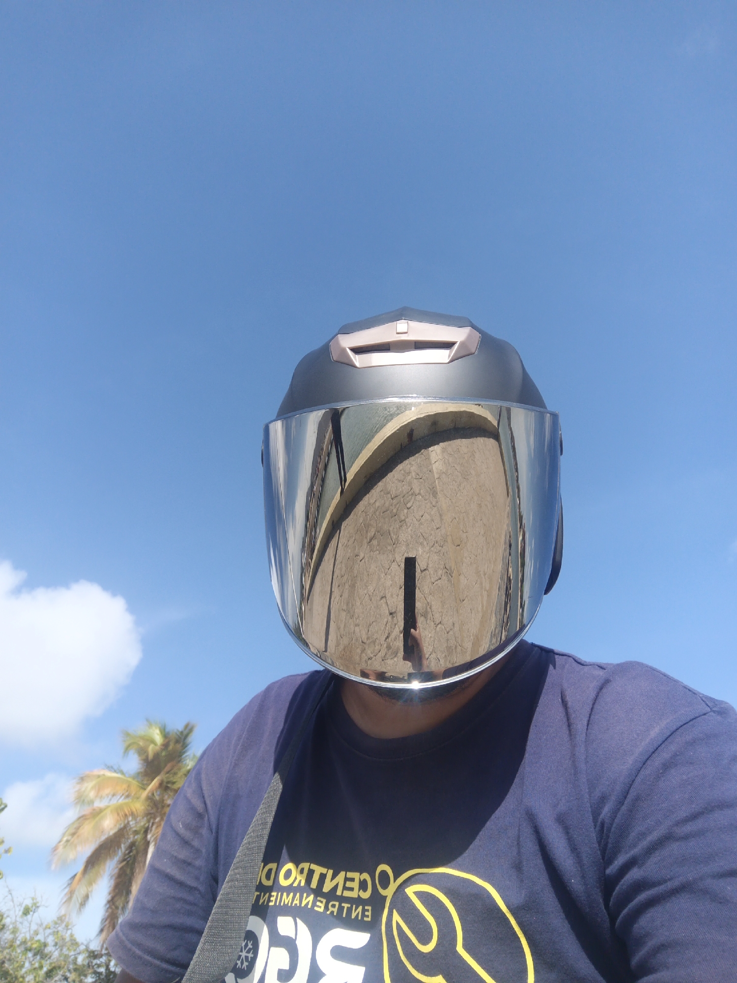A person wearing a motorcycle helmet with a reflective face shield takes a selfie outdoors. The face shield reflects the Castillo de San Carlos Borromeo, a historic fort, indicating that the photo was taken in or near Juan Griego, Venezuela. The person is wearing a dark blue t-shirt with a logo in yellow. Part of a palm tree and a bright blue sky with some clouds are visible in the background.