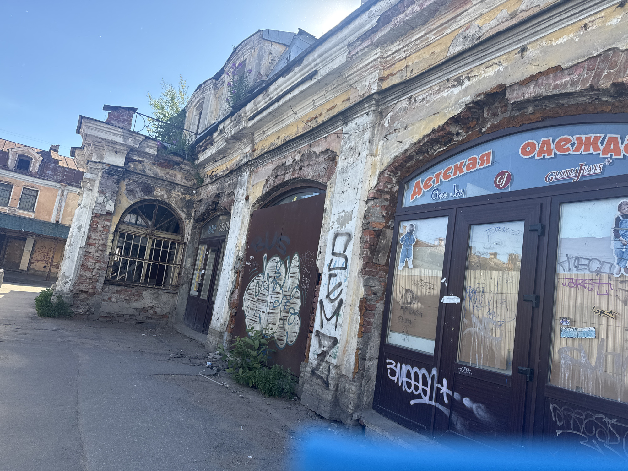 The image depicts an urban street scene in Saint Petersburg, Russia, featuring a dilapidated building with an arched storefront. The building exhibits significant deterioration, with peeling paint, exposed brickwork, and crumbling stucco. Graffiti adorns the building's facade, covering sections of the walls and doors. The storefront windows are also tagged with graffiti and some appear boarded up. An "Adskaya Odezhda" (which translates to "Children's Clothing") sign is visible above the storefront. A few sparse plants are growing in cracks and crevices around the base of the building. The street in front of the building is paved with asphalt and appears somewhat worn, with scattered debris. The sky is clear and blue, indicating a sunny day.