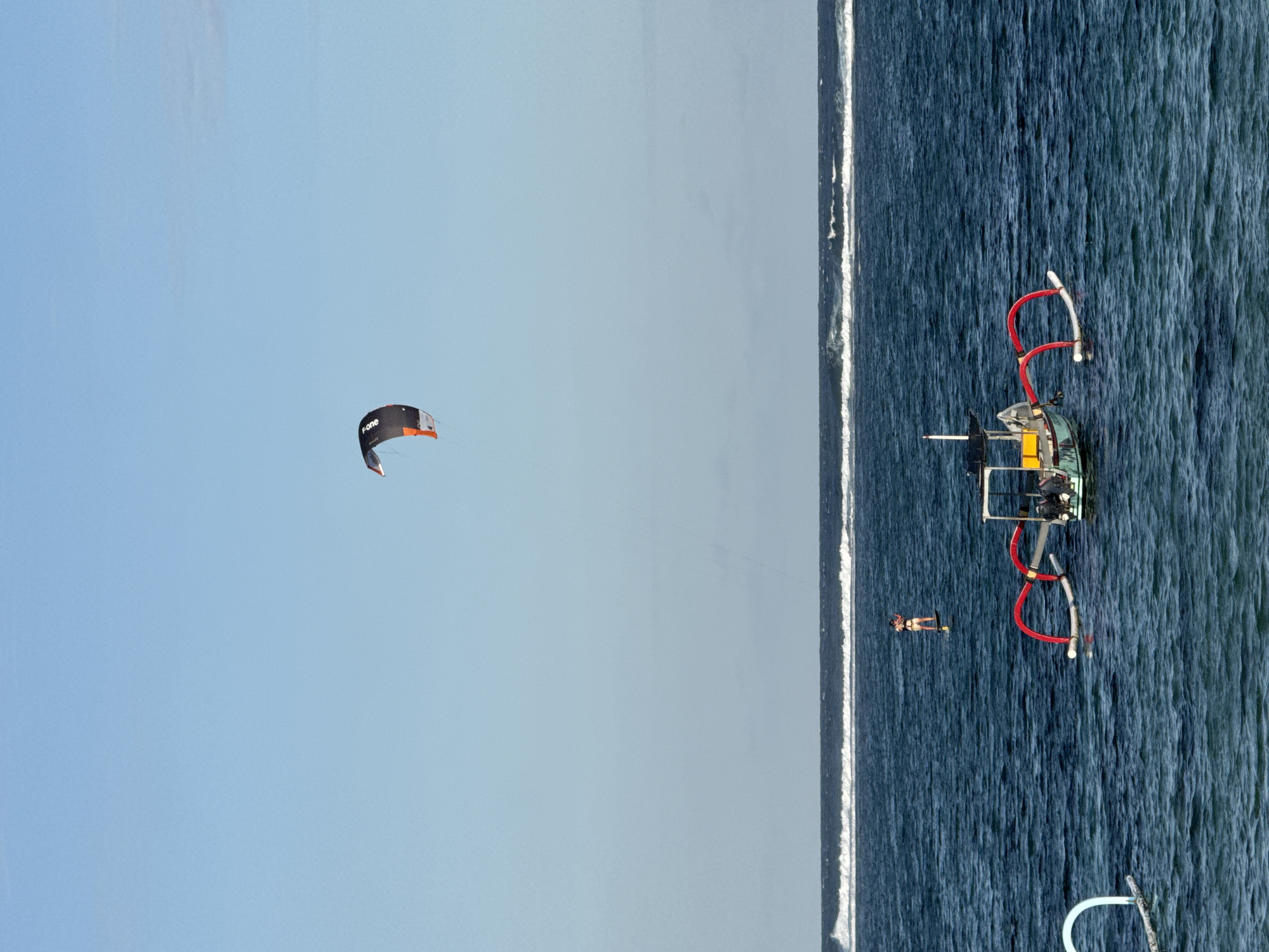 An aerial shot depicts a coastal scene, likely in Kuta Selatan, Indonesia, as evidenced by the watercraft. The view, taken at an angle, shows the sea meeting the sky. A person windsurfing is visible in the water near what appears to be the shoreline. A red watercraft is also present in the water. Above, a kite is flying in the clear blue sky, suggesting active wind sports in the area.