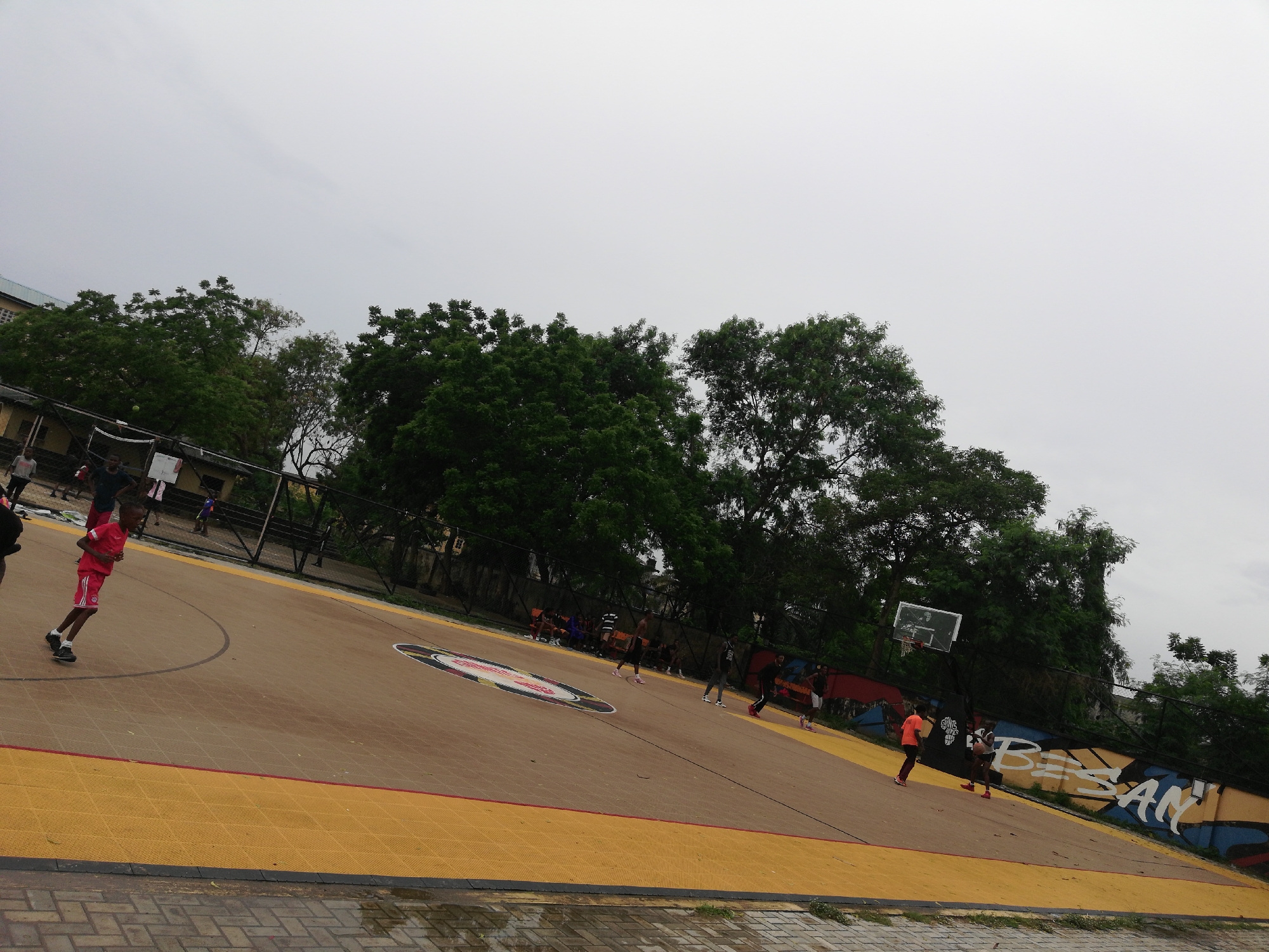 The image shows a basketball court in Lagos, Nigeria. The court has a brown and yellow surface with red lines. There is a group of people playing basketball. The environment is outdoors with trees surrounding the court. There is a mural on the wall next to the court that says "BESAN". The sky is overcast. The ground in front of the court looks wet.