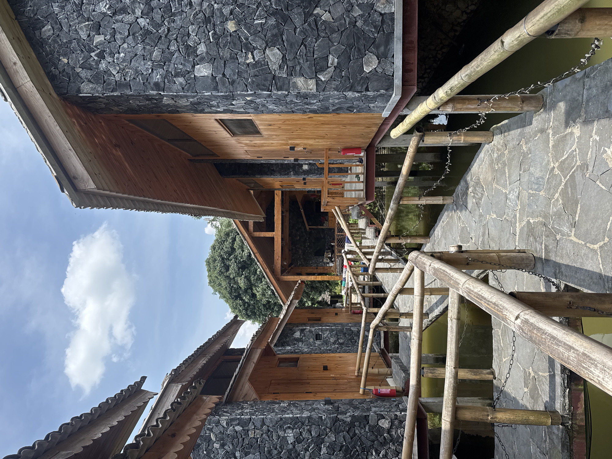 This media file captures a scenic view of traditional-style stilted buildings situated over calm, greenish water, likely part of a resort or homestay. The architecture features a blend of materials: the lower half of the buildings is constructed from dark, irregularly shaped stone masonry, providing a sturdy base, while the upper sections are made of warm-toned horizontal wooden planks. The roofs exhibit a distinctive scalloped or wavy edge, possibly made of timber or corrugated material, with a rustic aesthetic.
A narrow pathway, paved with irregular stone slabs, runs alongside the water, connecting the various structures. This path is bordered by a rustic handrail crafted from thick bamboo poles, which are secured with metal chains. Small, decorative string lights, possibly fairy lights, are visible strung along sections of the bamboo railing. In the background, lush green trees stand tall under a bright blue sky dotted with scattered white clouds, indicating clear daytime weather.
Red fire extinguishers are mounted on the wooden walls of some buildings, suggesting safety measures are in place. The scene appears devoid of people, contributing to a sense of tranquility and peacefulness. The use of natural materials like wood, stone, and bamboo, combined with the over-water setting, creates an ambiance typical of eco-friendly or traditional accommodations in a natural landscape. No discernible text is visible in the image.