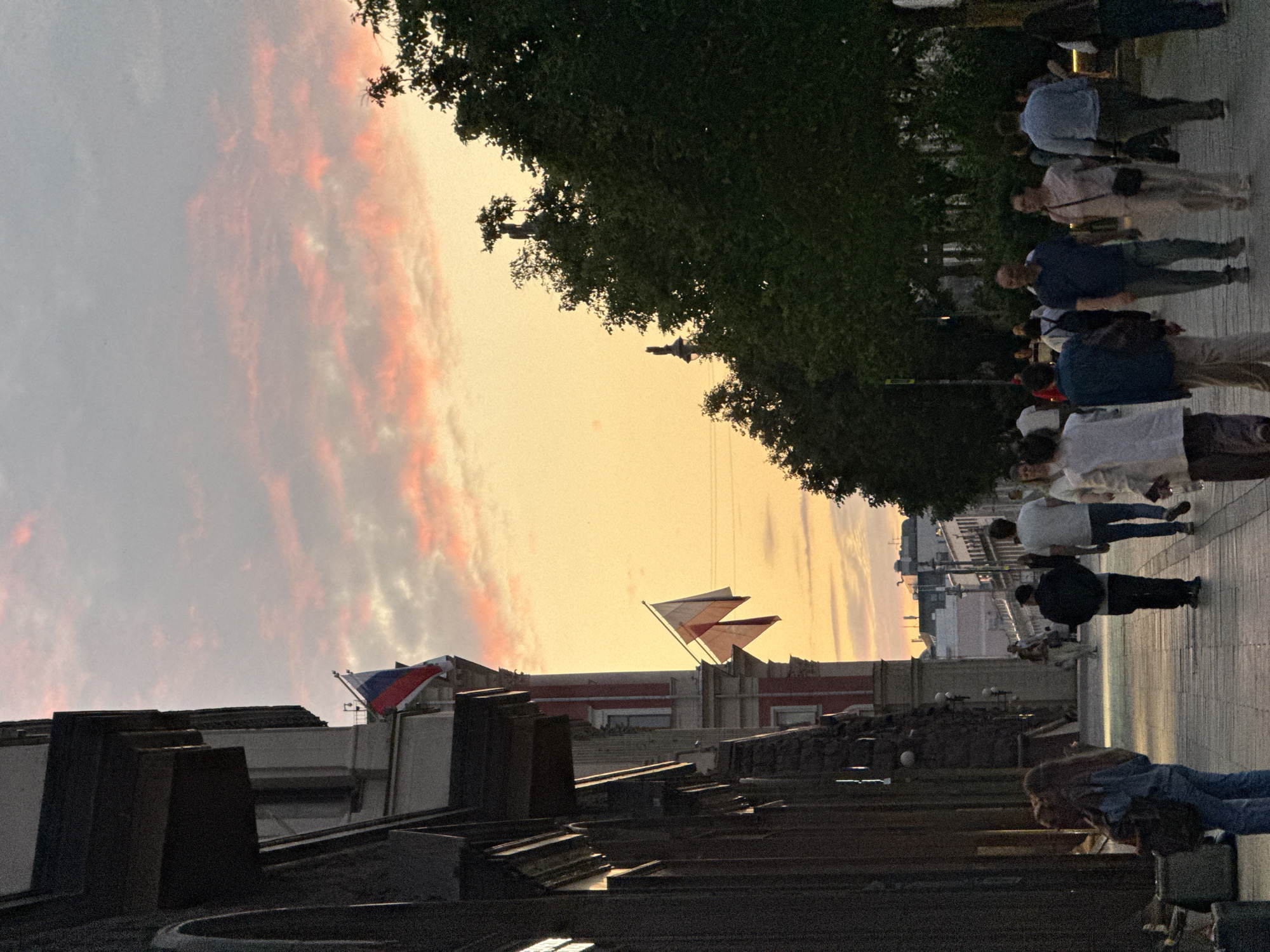 This is an eye-level, medium-shot, vertical photograph taken in Moscow, Russia, during twilight. The scene captures a street with pedestrians walking on a sidewalk beside a building and trees.
On the left side of the frame, a tall, dark building with architectural detailing is visible. Two Russian flags are displayed on the building: one near the top with its red and blue stripes clearly visible against the sky, and another lower down, also displaying the Russian tricolor.
The sky is a prominent feature, showcasing a gradient of colors from yellow at the horizon to pink and gray clouds higher up.
On the right side of the frame, large green trees create a natural backdrop. In the distance, the silhouettes of more buildings and streetlights can be seen.
A diverse group of people are walking along the sidewalk. They are dressed in casual clothing, and their activities suggest a leisurely stroll. Some are walking alone, while others are in small groups. Their faces are not clearly discernible due to the lighting conditions and distance. The overall atmosphere appears calm and serene, typical of an early evening in a city.