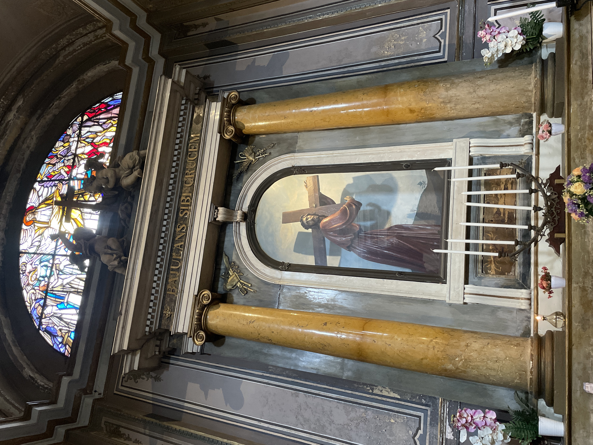 The image shows an ornate religious altar inside what appears to be a church in Milan, Italy. A stained-glass window featuring figures is on the left, next to the altar which displays a painting of Jesus carrying the cross. The painting is flanked by faux marble columns with decorative capitals. Floral arrangements and candles adorn the altar. The inscription "BAJULANS SIBI CRUCEM" is visible on the side of the altar.
