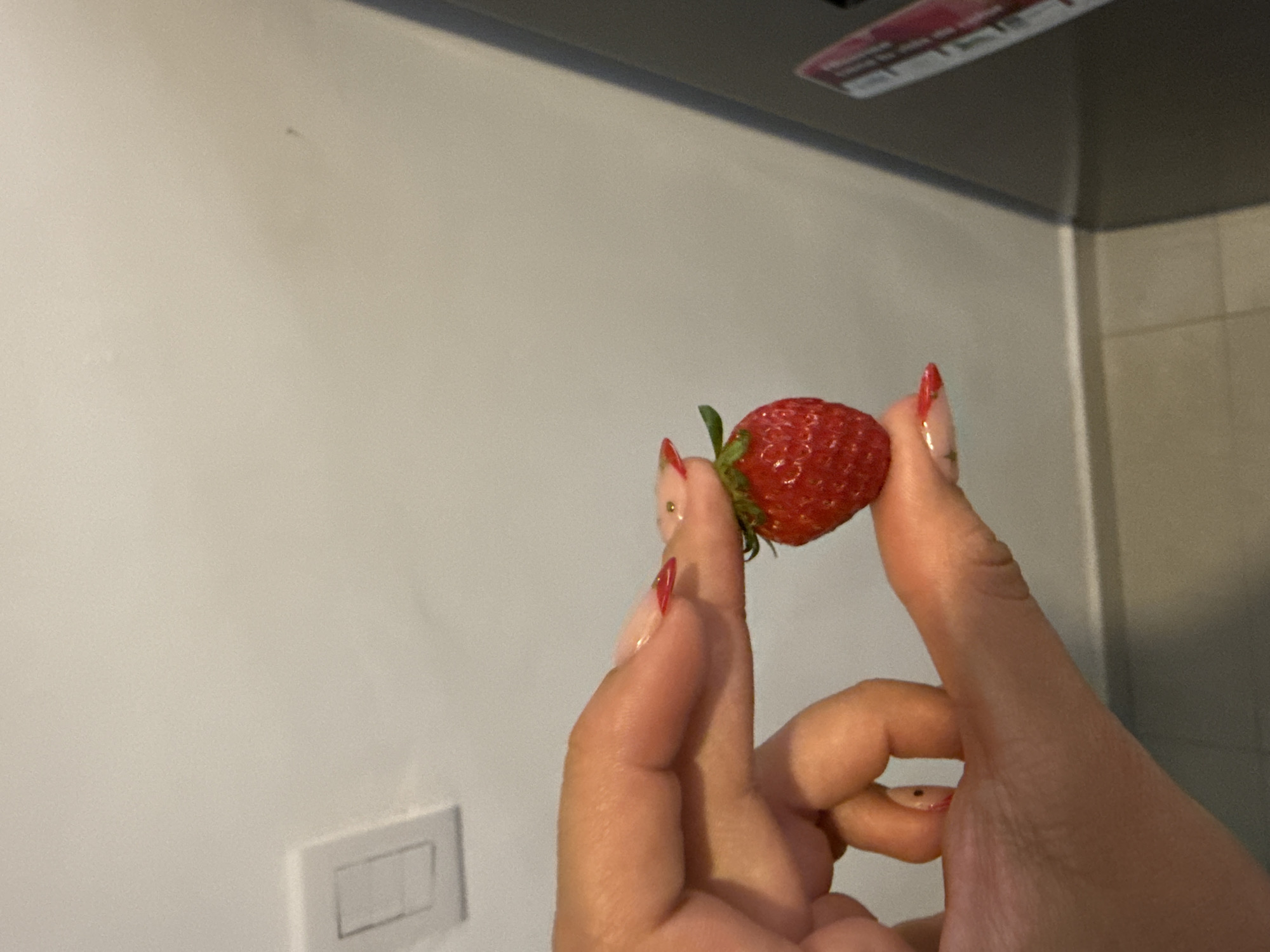 The image displays a close-up of a hand delicately holding a single, ripe strawberry. The strawberry is vibrant red with its green stem (calyx) still attached, positioned between the person's thumb and index finger. The nails on the visible fingers are manicured with a clear or light-colored base and distinct red tips, characteristic of a French manicure style.
The background primarily consists of a plain, light-colored wall, likely off-white or beige. In the lower-left portion of the frame, a white light switch featuring two rectangular buttons is partially visible against the wall. Towards the top of the image, a dark grey or black overhead object can be seen, which appears to be an appliance such as a range hood or a cabinet. The underside of this object shows what looks like a label or sticker with some reddish and white elements, but any text present is too blurry to be read.
The setting is indoors, with artificial lighting illuminating the scene. The image provides no specific indicators for the time of day or weather conditions. Based on the provided context, the location of this scene is Puebla, Mexico. No emotions are discernible from the partial view of the hand.