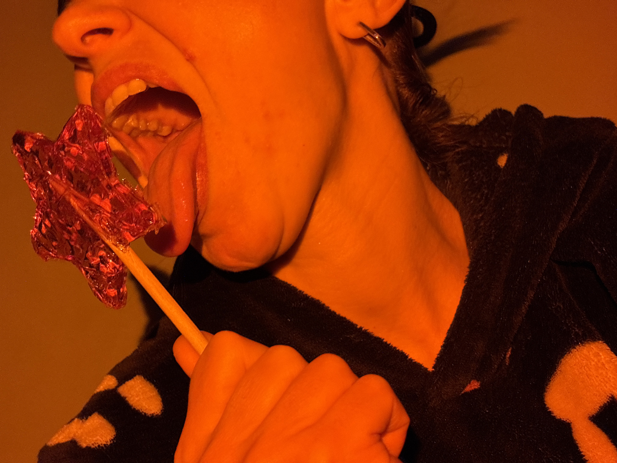 A person, likely female, is depicted with their mouth open, tongue extended, and a star-shaped lollipop partially inside. They are wearing a dark hooded garment with a spotted pattern. The lighting appears to be predominantly orange. The location is not discernible from the image.
