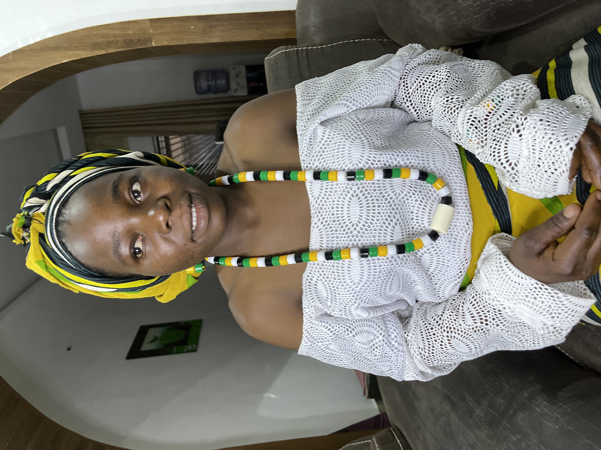 A medium shot of a dark-skinned woman wearing a yellow, green, black, and white head wrap. She has a necklace of the same colors. She is wearing a white lace shirt with long sleeves. She is sitting in a brown chair. The background is a light-colored wall with a framed picture. There is an archway in the upper left corner of the frame. The location is likely indoors, possibly in a home in Jos, Nigeria.