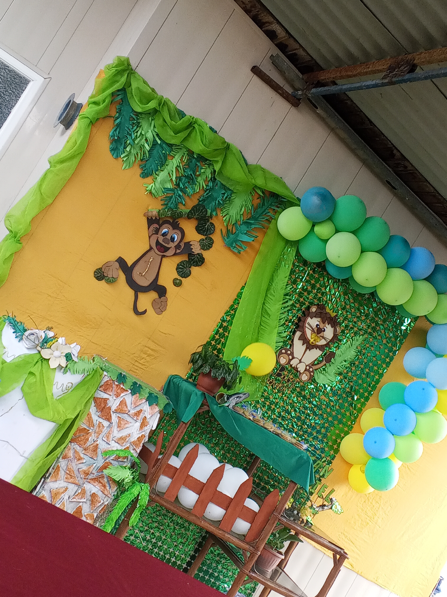 A decorated jungle-themed setup is visible in Altagracia, Venezuela. A yellow backdrop is adorned with green fabric, faux foliage, and a cut-out monkey. Adjacent to this, a green sparkly background features a lion cut-out and an arch of green, blue, and yellow balloons. Below, a small wooden structure with white cushion is decorated with greenery.