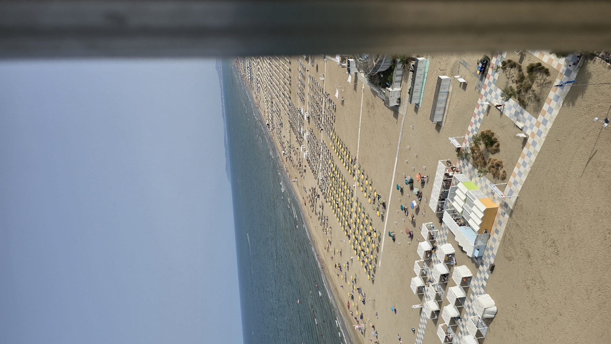 The image displays a panoramic view of a crowded beach in Rimini, Italy, likely during the daytime. The beach is lined with rows of yellow and white beach umbrellas and lounge chairs, with many people relaxing under them. The sea, a calm blue, stretches to the horizon, with a few boats visible. The shoreline is busy with sunbathers and swimmers. Closer to the foreground, there are beachside facilities including white cabanas, colorful small buildings, and paved walkways with checkered patterns. A few people are walking on the beach and along the pathways. The overall scene depicts a vibrant summer day at the beach, characteristic of Rimini's popular seaside resorts.