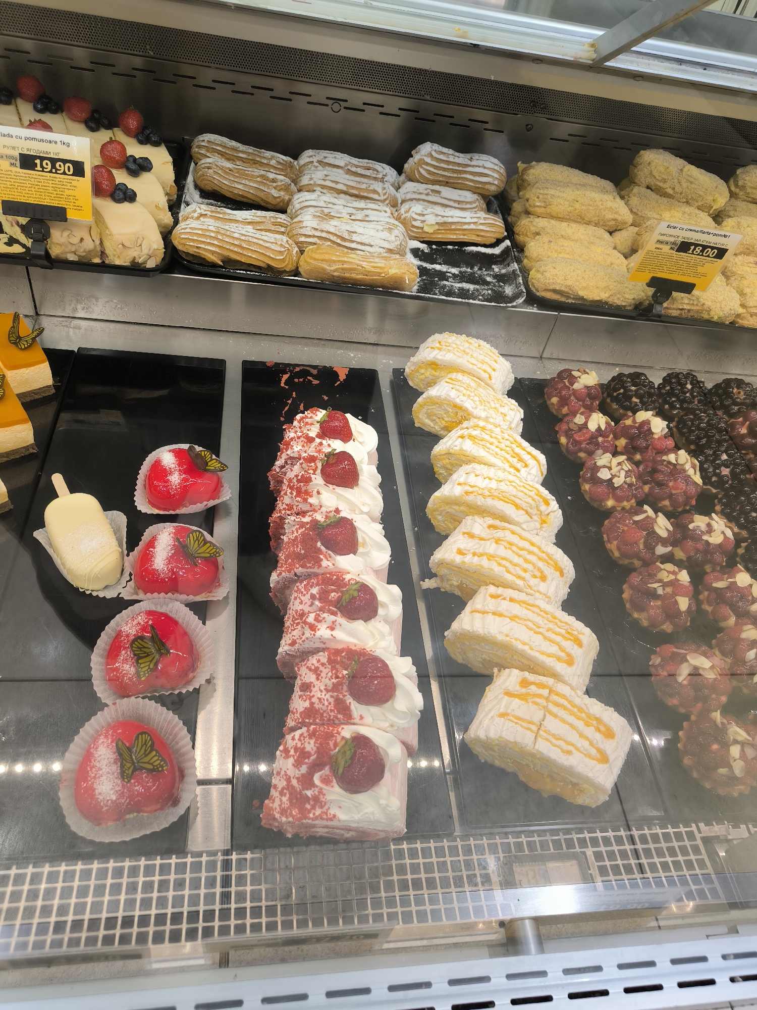 The image displays a refrigerated display case filled with various pastries, likely in a bakery or supermarket in Chișinău, Moldova. The pastries are arranged on black trays and include eclairs, layered cakes with frosting, fruit tarts, and individual heart-shaped cakes topped with butterflies and shredded coconut. Some of the pastries are accompanied by price tags, such as one for "Ecler cu cremai flart-pomlet" priced at 18.00. Another tag indicates a price of 19.90 for a cake labeled "lada cu pomusoare". The environment appears clean and well-lit, typical of a commercial food establishment. No people are visible in the frame.