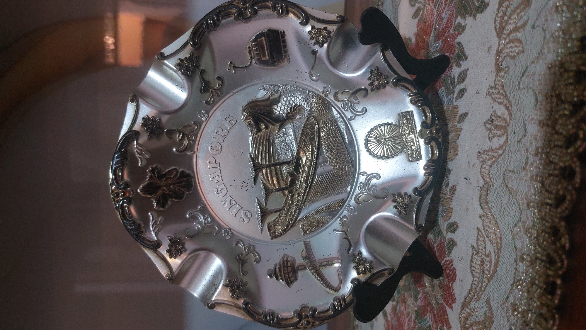 The image shows a silver-colored decorative plate, likely a souvenir, depicting various landmarks of Singapore. The plate is ornately decorated with floral motifs and has a scalloped edge. In the center, the word "SINGAPORE" is embossed. Prominently featured are the Merlion statue, Marina Bay Sands, the Singapore Flyer, and the Singapore Cable Car. The plate is displayed on a black stand, placed on a surface covered with a patterned cloth. The background is dark and appears to be indoors. The lighting is dim, with some reflections visible on the plate's surface. There are no people visible in the image. This decorative plate is likely a tourist item, and its presence in the city of Medan, Indonesia, would be due to it being a souvenir or a collectible item.