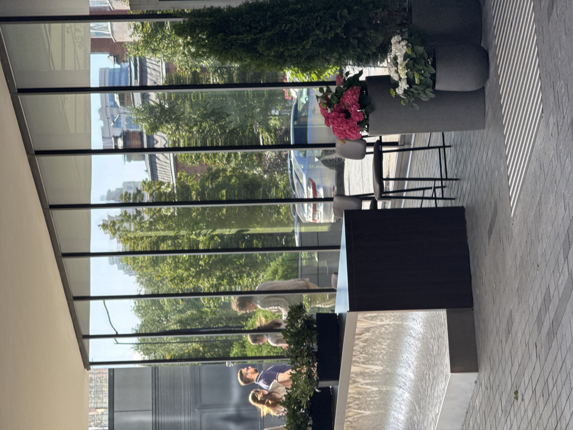 The image depicts a sunlit outdoor patio area with large glass windows reflecting trees and buildings. In the foreground, two planters hold vibrant pink and white flowers. A dark-colored table and chairs are visible, suggesting an outdoor dining or seating area.
Through the glass, reflections reveal a street scene with trees, cars, and buildings. People are visible in the reflections. One person appears to be bending down, while another is walking. Further down, two people are standing together, one with blonde hair and the other wearing glasses and a blue and white striped shirt.
The lighting suggests it is daytime, with strong sunlight creating shadows on the tiled ground. The scene appears to be a modern urban setting. The reflection of trees indicates a pleasant weather day. The overall mood is relaxed and casual. There is no visible text in the image.