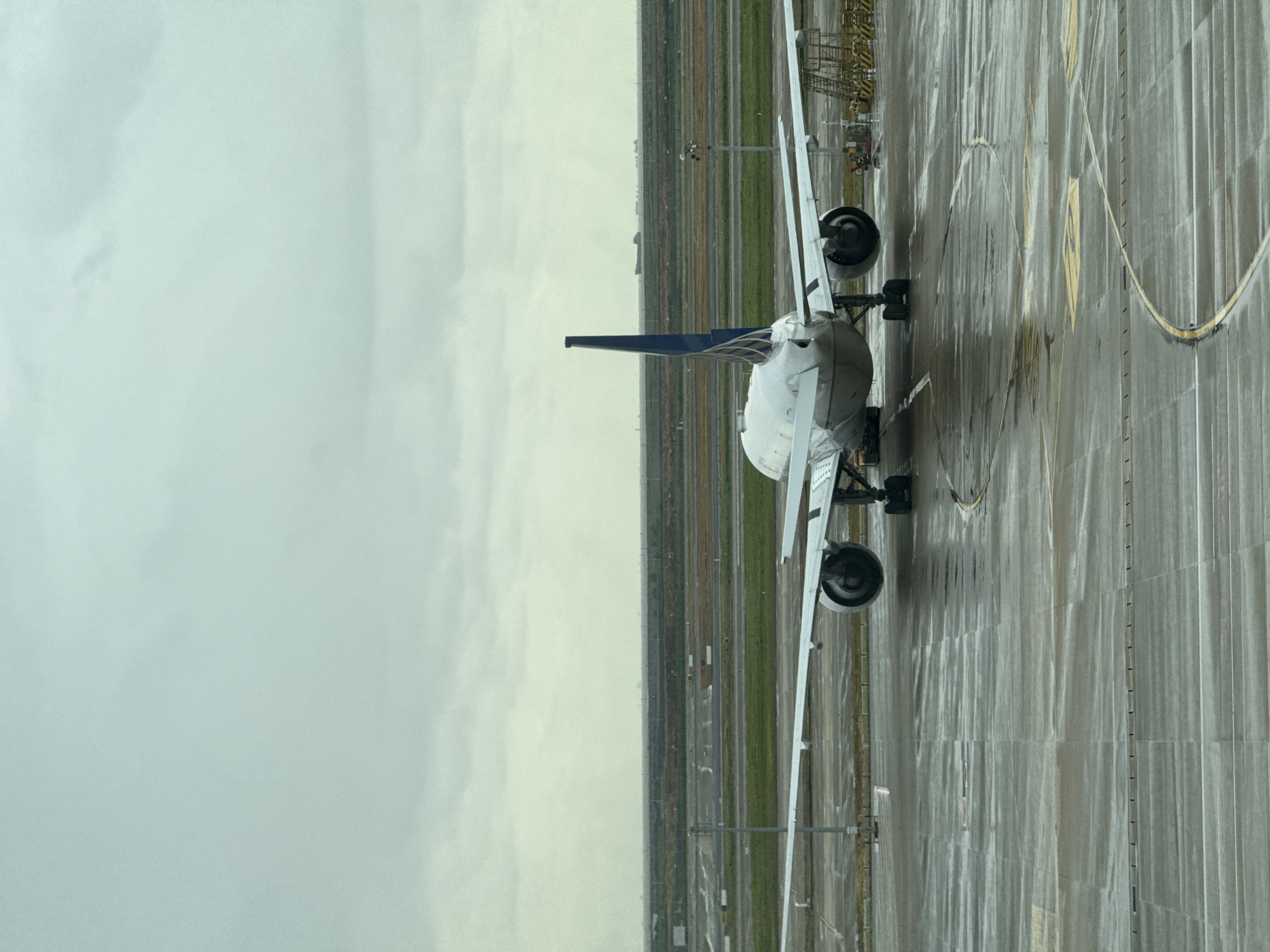 A wide shot shows the rear view of a large commercial airplane parked on a wet tarmac at Pudong International Airport in Pudong, China. The plane is a wide-body jet with two engines under its wings, and its tail is a dark blue with a white logo. The tarmac is wet and reflective, suggesting it has recently rained, and there are yellow lines marking parking or taxiing areas. In the background, there are rows of buildings and greenery stretching to the horizon, with a cloudy, overcast sky overhead. A few distant ships can be seen on what appears to be a body of water in the far background. There are no people visible in the image. The scene is one of a typical airport environment, with the focus on the stationary aircraft.