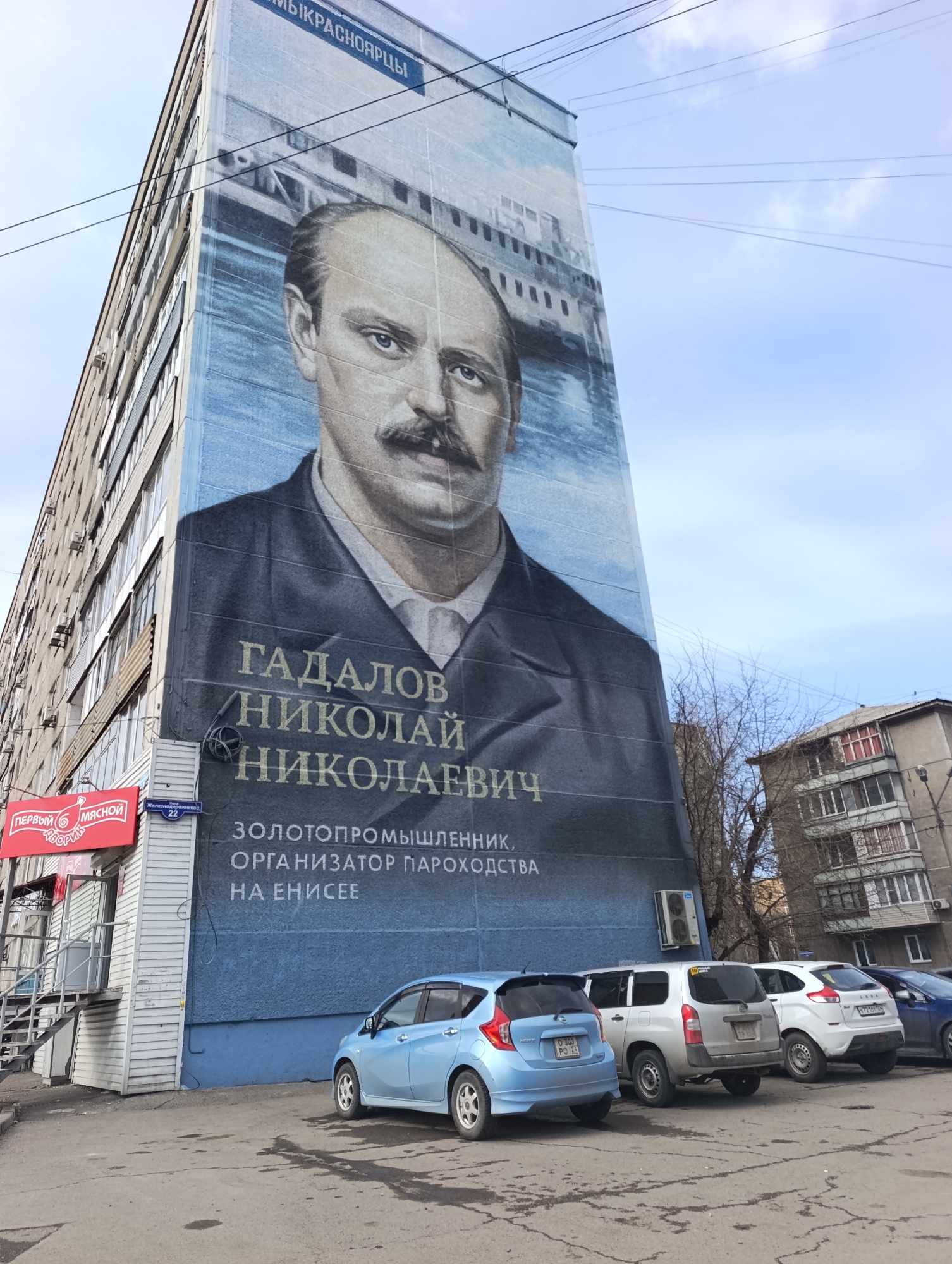 This media file captures an urban street scene in Krasnoyarsk, Russia, dominated by a large mural covering the entire side of a multi-story residential building. The mural features a prominent portrait of a serious-looking man with a mustache, identified by the accompanying Russian text as "ГАДАЛОВ НИКОЛАЙ НИКОЛАЕВИЧ" (Gadalov Nikolai Nikolaevich). Further text describes him as a "ЗОЛОТОПРОМЫШЛЕННИК, ОРГАНИЗАТОР ПАРОХОДСТВА НА ЕНИСEE" (Gold industrialist, organizer of steamship navigation on the Yenisei). The top of the mural also shows "КРАСНОЯРЦЫ" (Krasnoyartsy), referring to the people of Krasnoyarsk.
In front of the mural-adorned building, several cars are parked on a paved lot, including a light blue Nissan hatchback (license plate O 300 PO 24), a silver compact wagon, and a white crossover (license plate H 721 OH 24). The "24" on the license plates denotes Krasnoyarsk Krai.
On the ground floor of the mural building, a commercial establishment is visible with a red sign that reads "ПЕРВЫЙ МЯСНОЙ ФАВОРИТ" (Pervy Myasnoy Favorit - First Meat Favorite). A street sign near the entrance indicates "Железнодорожников 22" (Zheleznodorozhnikov 22 - Railroad Workers Street 22).
In the background to the right, another multi-story residential building and bare trees suggest a cooler season. Overhead power lines stretch across the clear, bright sky with scattered clouds. The scene is static, depicting a typical daytime urban landscape.