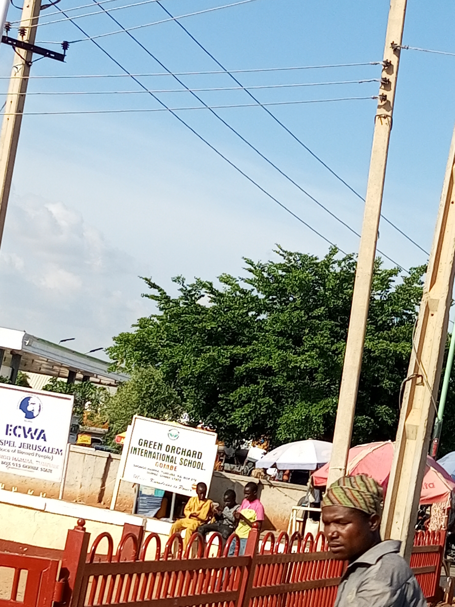 This media file captures an outdoor scene in Tumfure, Nigeria, likely during the daytime under a clear blue sky with some scattered clouds. The foreground features a decorative red fence and a man wearing a headwrap and a grey shirt, looking to the right. Behind him, several people are visible, some seated and others standing, near a market or roadside stalls with umbrellas providing shade.
Two signs are prominently displayed. One reads "ECWA GOSPEL JERUSALEM" with some additional text below, suggesting a religious institution. The second sign announces "GREEN ORCHARD INTERNATIONAL SCHOOL, GOMBE," also with further details.
The background is dominated by lush green trees and overhead power lines supported by utility poles, indicating an urban or semi-urban environment. The overall atmosphere appears to be that of a bustling community with commercial activity and educational institutions present. The sunlight suggests it is daytime, and the clear sky points to fair weather.