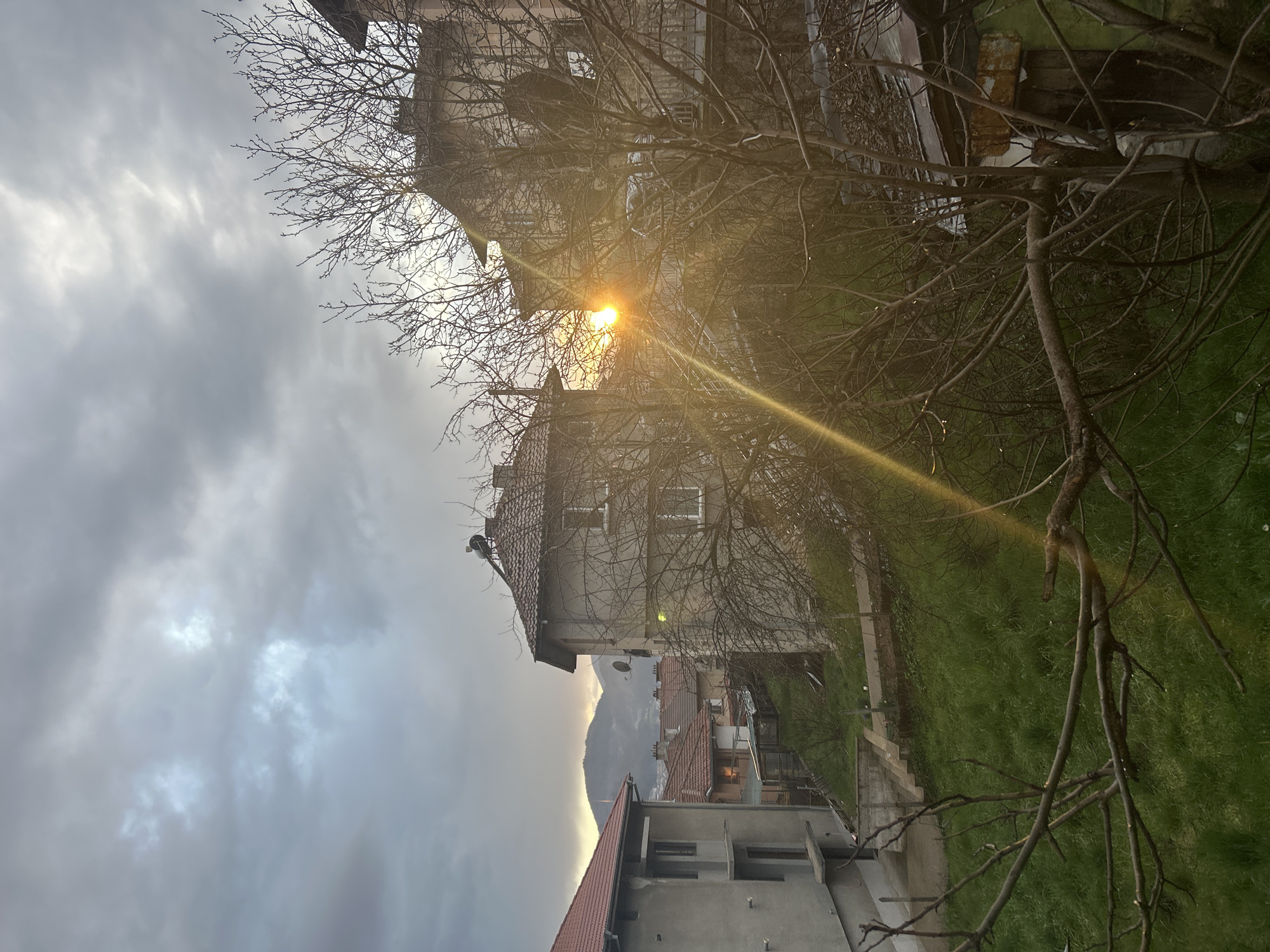 The image depicts an urban landscape with buildings, bare trees, and a cloudy sky. Sunlight is partially obscured by the buildings and branches, creating a lens flare. In the background, a mountain is visible. The buildings appear to be residential, with varying architectural styles. The scene suggests an early morning or late afternoon setting. The photograph does not explicitly confirm the location in Zlatograd, Bulgaria, but the scene is consistent with urban areas in that region.