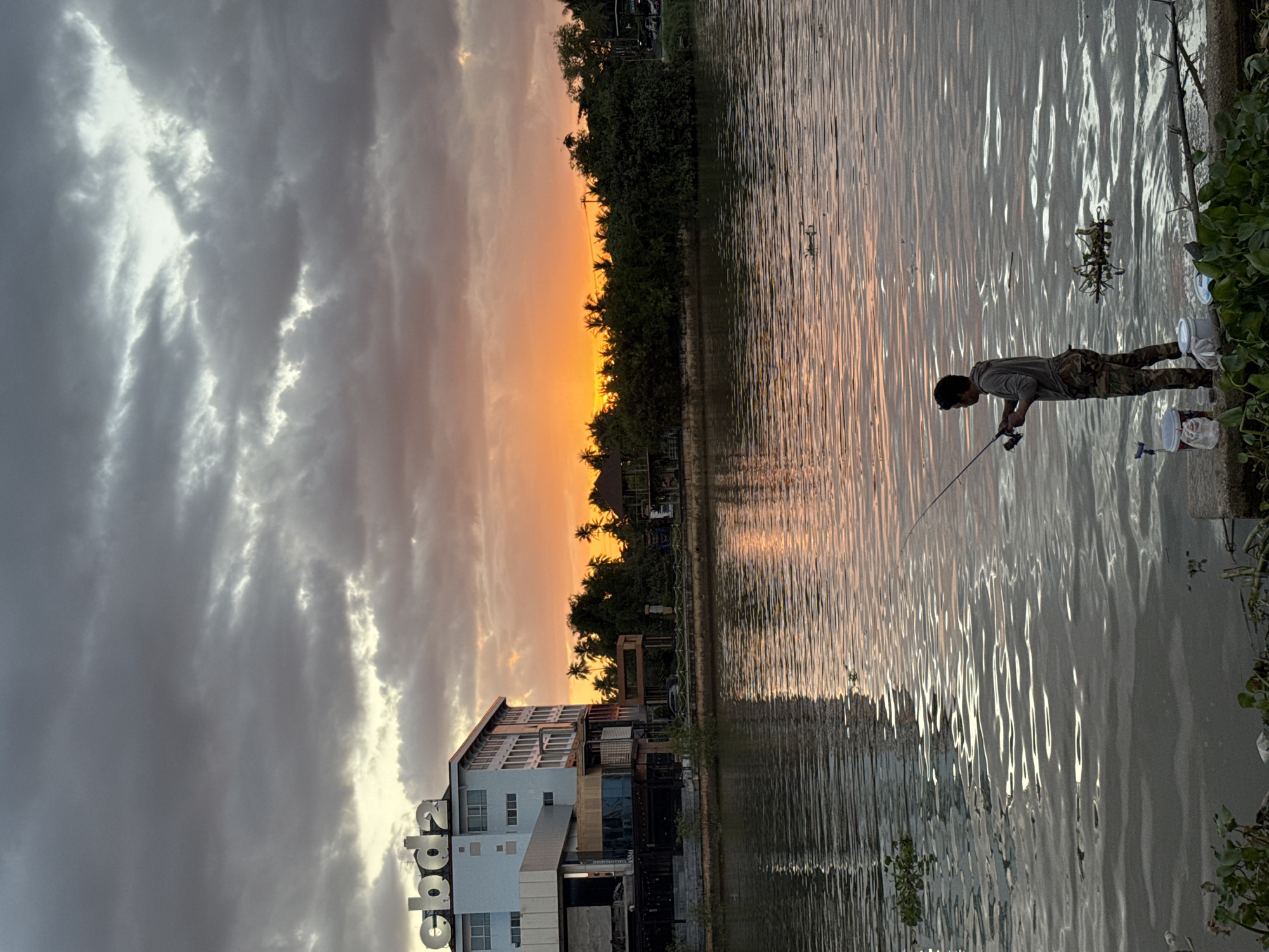 The image depicts a sunset scene on a riverbank in Surat Thani City Municipality, Thailand. A man is standing on a concrete embankment, holding a fishing rod and fishing in the river. He is wearing camouflage pants and a grey hoodie. Two buckets are placed on the ground near him. The river reflects the warm colors of the sunset. In the background, a multi-story building with the letters "cbd2" on its facade is visible on the left. Across the river, a dense line of trees and vegetation stretches along the bank, with palm trees interspersed. The sky is filled with dramatic clouds, illuminated by the setting sun, creating a vibrant orange and yellow glow.