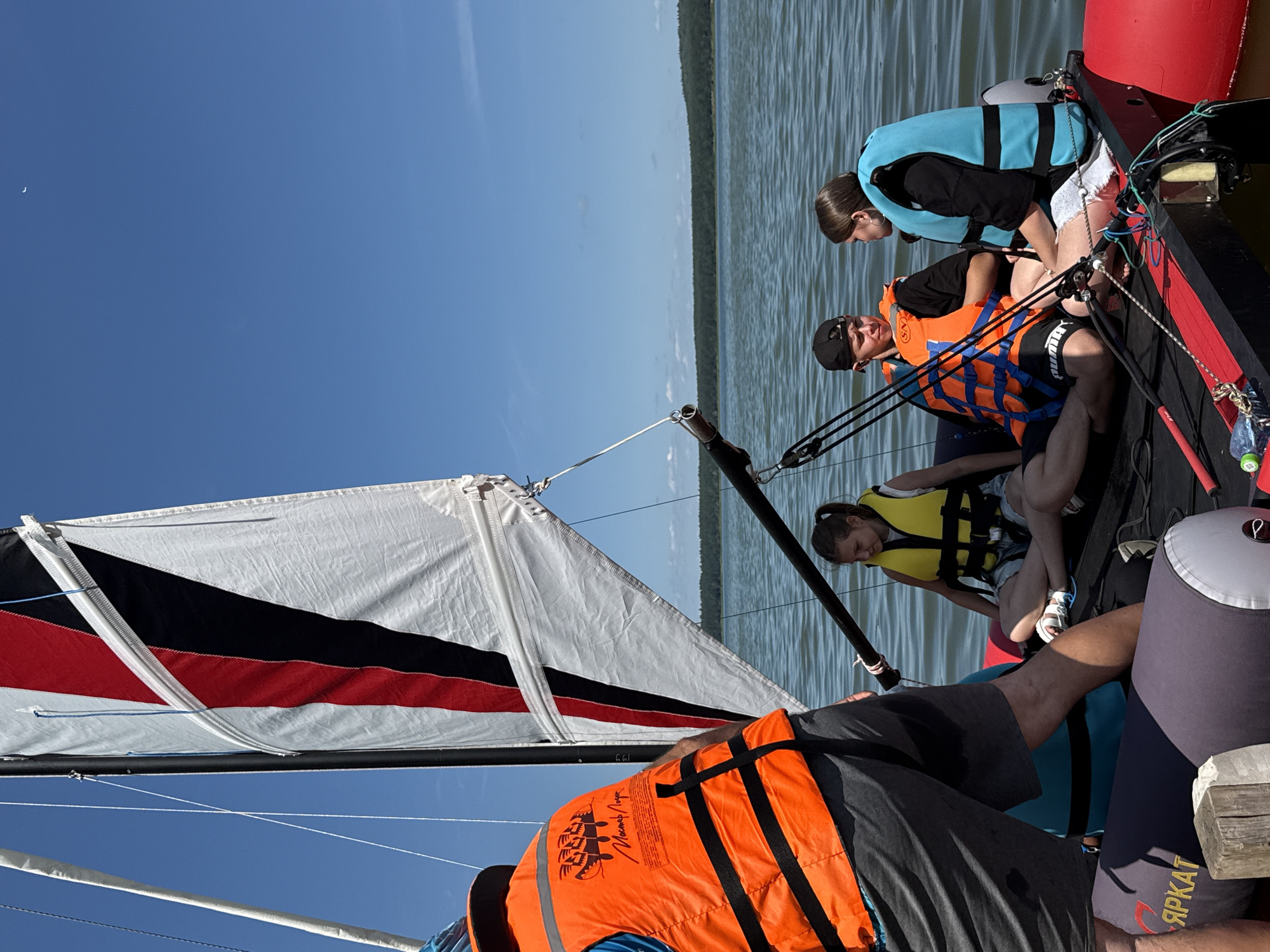 Four people, three of whom are wearing life vests, are seated on a sailboat on a body of water. The sailboat has a large sail with red, black, and white sections. The people are in the foreground, with a tree line in the background. The sky is clear and blue. The scene is set on Lake Svetloe in Votkinsk, Russia.
