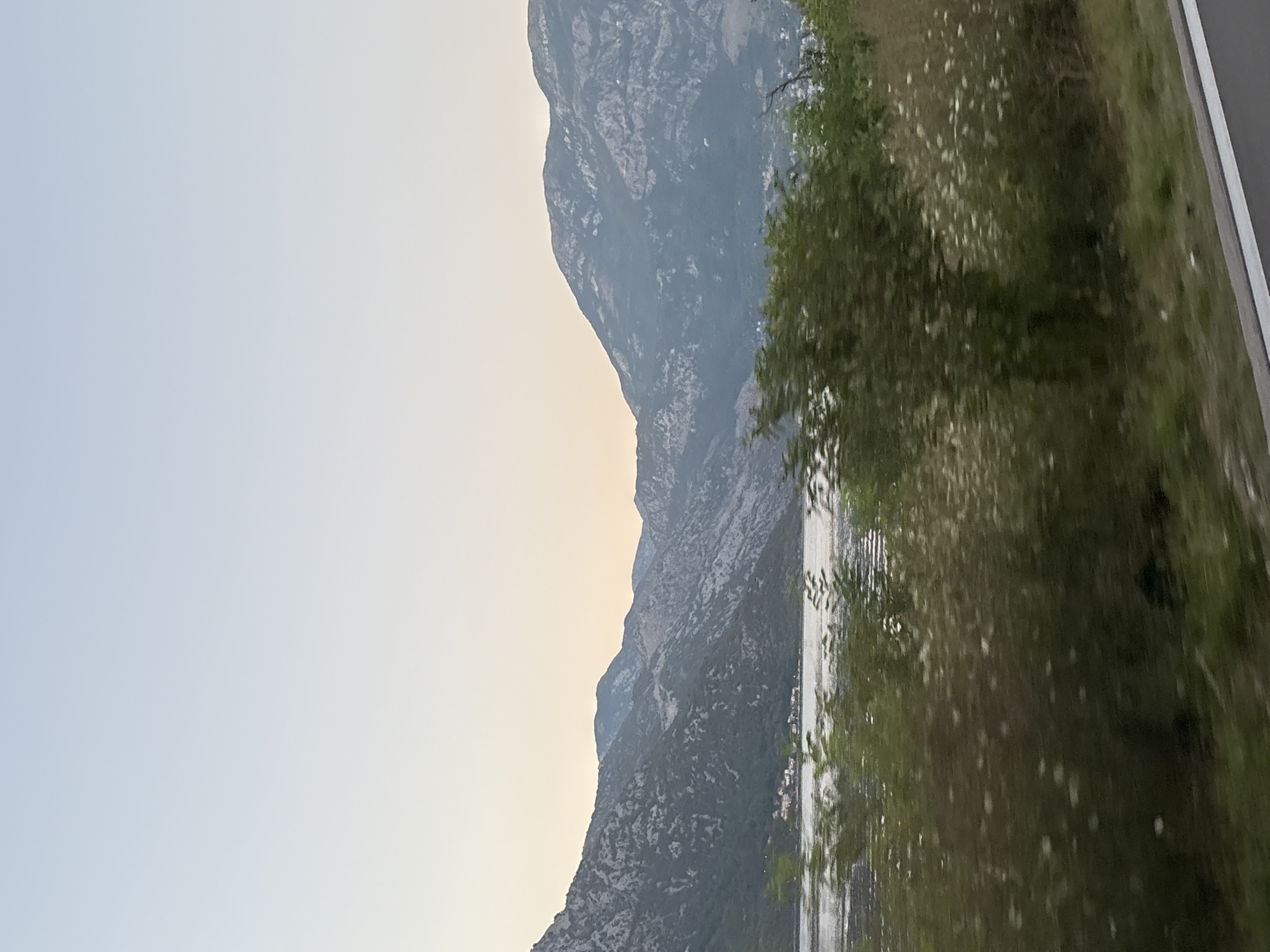 The image depicts a scenic view of the Bay of Kotor in Kostanjica, Montenegro. The foreground features dense green foliage, including trees and bushes with small white flowers, bordering a road with a white line. Beyond the vegetation lies the calm, reflective water of the bay, with ripples suggesting a gentle movement. In the mid-ground, a small coastal settlement with white buildings is visible nestled against the steep, mountainous terrain. The background is dominated by imposing, rocky mountains, characterized by their rugged textures and varying shades of gray and blue, suggesting a vast and dramatic landscape. The sky above is clear and pale, transitioning from a light blue at the top to a softer, warmer hue near the horizon, indicating either early morning or late evening light. The overall impression is one of natural beauty and tranquility, with the human settlement appearing small and integrated into the grand scale of the natural environment.