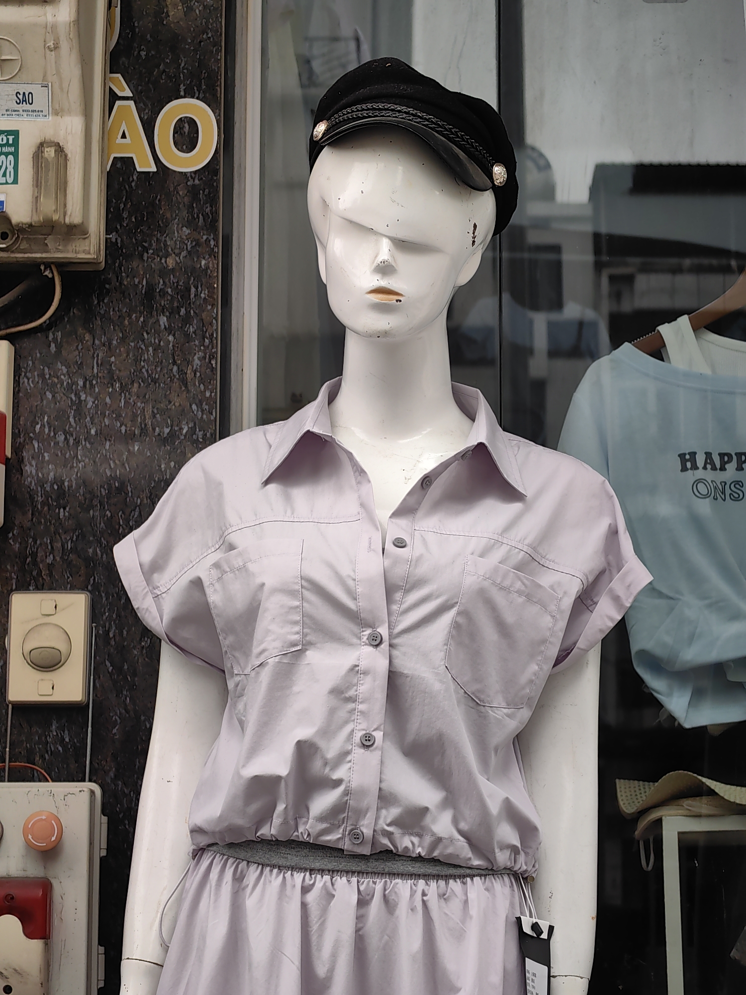 A medium shot captures a mannequin wearing a black cap and a light purple outfit, likely displayed in a store window. The location is Bai Chay Ward, Vietnam, suggested by the Vietnamese characters on a nearby electrical box labeled "SÃO" and "ÃO".
The mannequin is white with an indistinct face, wearing a black peaked cap with a gold button. The mannequin is wearing a purple collared shirt with short cuffed sleeves, two breast pockets, and buttons down the front. The shirt is tucked into a matching purple skirt with a drawstring waist. A tag hangs from the skirt.
To the left of the mannequin is an electrical panel with visible components. To the right, clothing items on a hanger are partially visible in the reflection on the window.