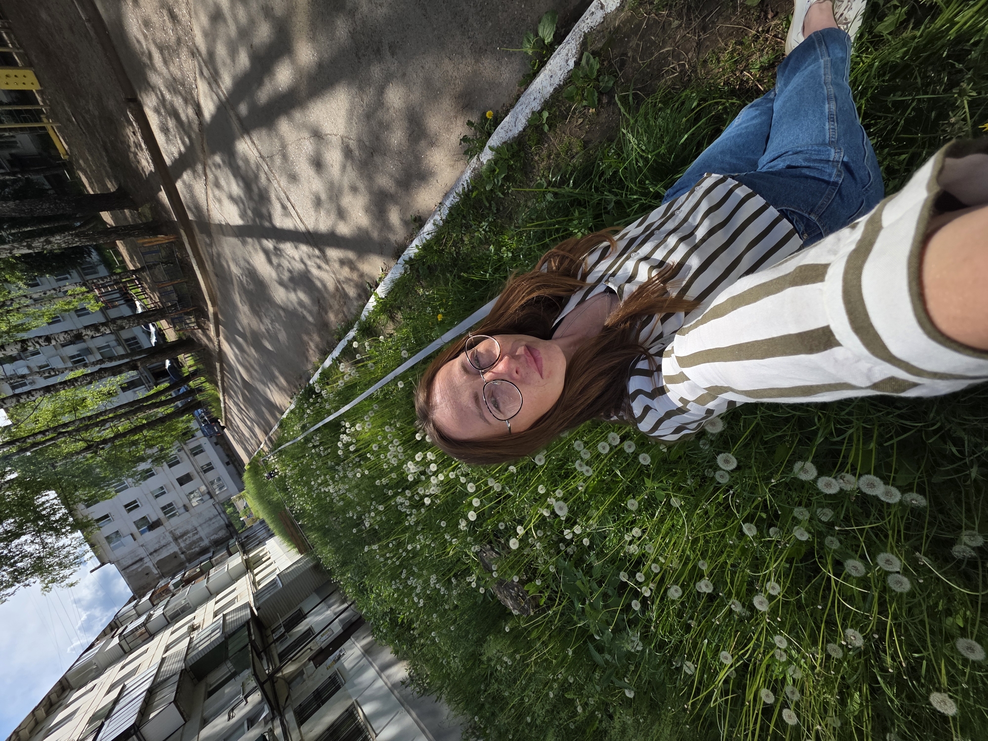 A young woman lies on her back in a dandelion-filled grassy area in Chaikovsky, Russia, taking a selfie. She wears glasses, a striped shirt, and jeans, and is positioned near the edge of the grass with a sidewalk and buildings in the background. Shadows from trees are visible on the sidewalk.