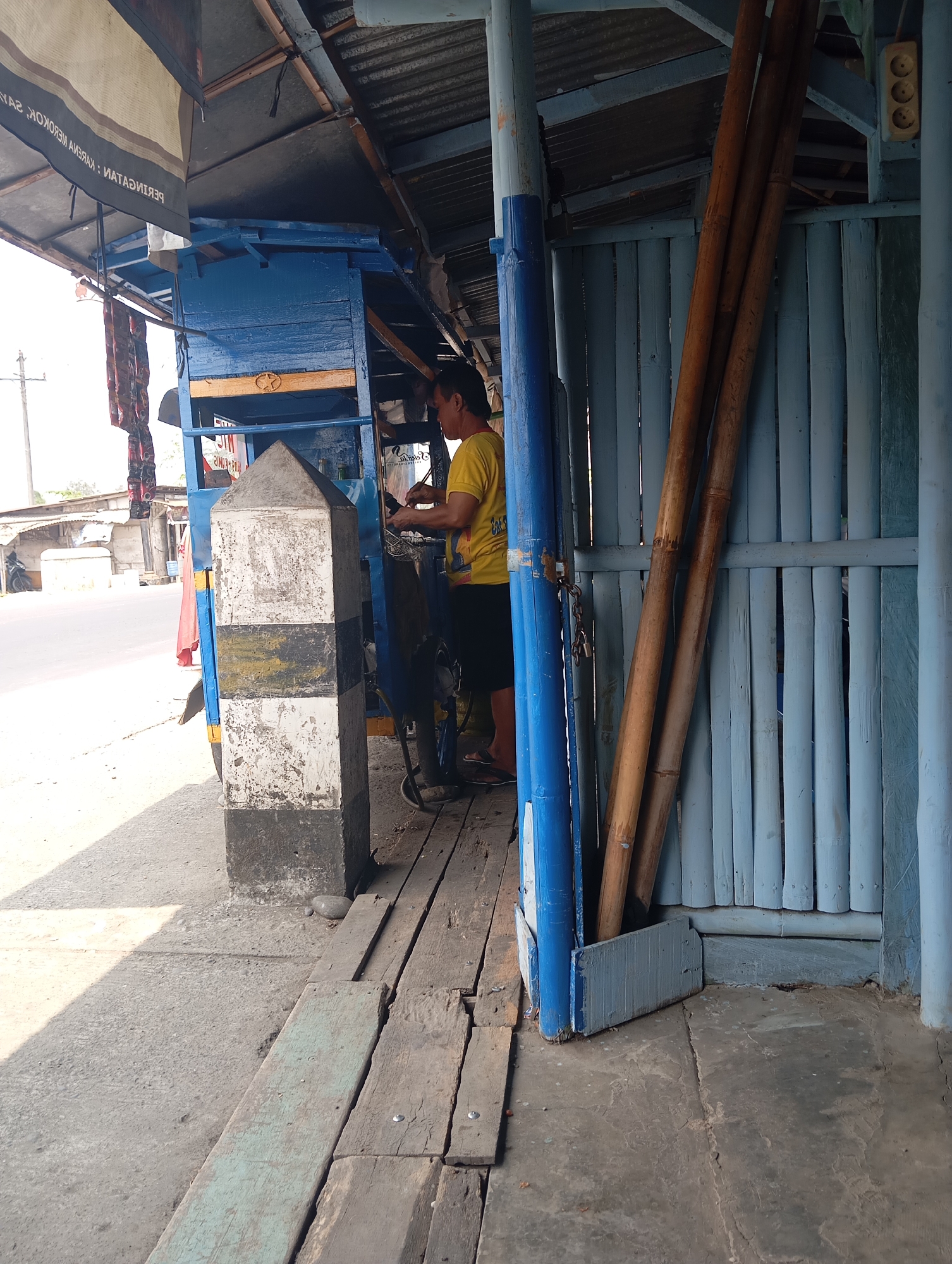The image shows a roadside scene in Sokaraja Wetan, Indonesia, with a man standing near a blue and yellow cart. The cart is partly covered by a corrugated metal roof, supported by wooden beams and a blue pole. In the foreground, a striped concrete marker, painted black, white, and yellow, stands on the sidewalk. To the right of the marker, several long bamboo poles are leaning against a light blue, slatted wall. The man, wearing a yellow t-shirt and dark shorts, is seen from the back, facing the cart and appearing to be working on something. A chain is visible hanging from a blue pole. The ground is a mix of concrete and wooden planks. In the background, a street with a few buildings and a motorcycle is visible, suggesting an urban or semi-urban setting.