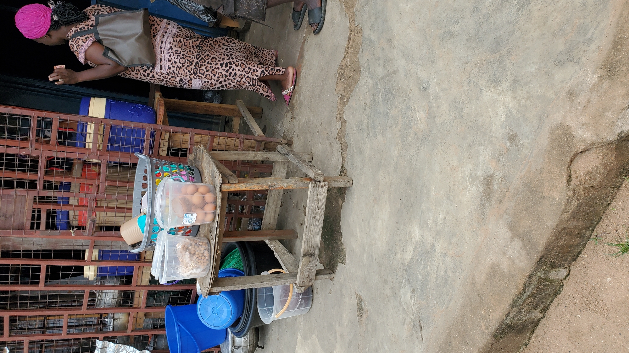 A woman wearing a pink headscarf and a leopard-print dress stands in front of a market stall in Jalingo, Nigeria. She holds a small object in her right hand. A large grey woven basket is visible, containing a tan cylindrical object and a blue and tan object. Two clear plastic containers are placed on a wooden stool in front of the basket; one contains what appears to be dried legumes or nuts, and the other contains a dozen brown eggs. Behind the stool, several blue and black plastic buckets and containers are stacked. The stall is covered by a metal grate structure. The ground is concrete, with some cracks and discoloration.