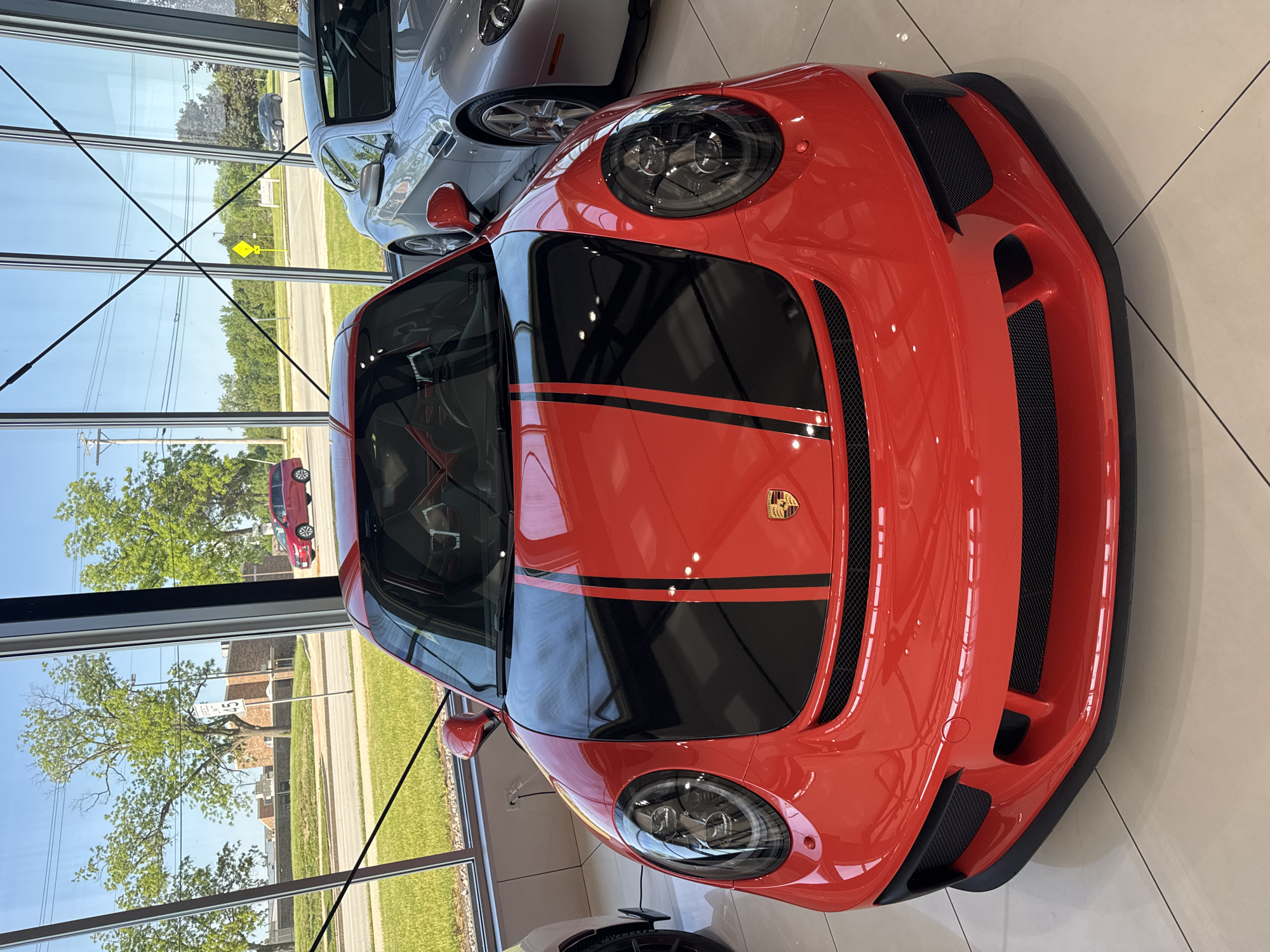 The image shows the front of a red Porsche car with black racing stripes, positioned inside a showroom or dealership. Another silver car is partially visible behind it. The showroom has large windows, and the reflection in the windows reveals a view of a street outside, indicating the presence of trees, buildings, and possibly a road. The location is Naperville, United States of America, as it is visible in the reflection of a street sign. The showroom floor appears to be tiled. There are no people visible in the image. The lighting is bright, suggesting it is daytime. The angle is from above and slightly to the side of the car.