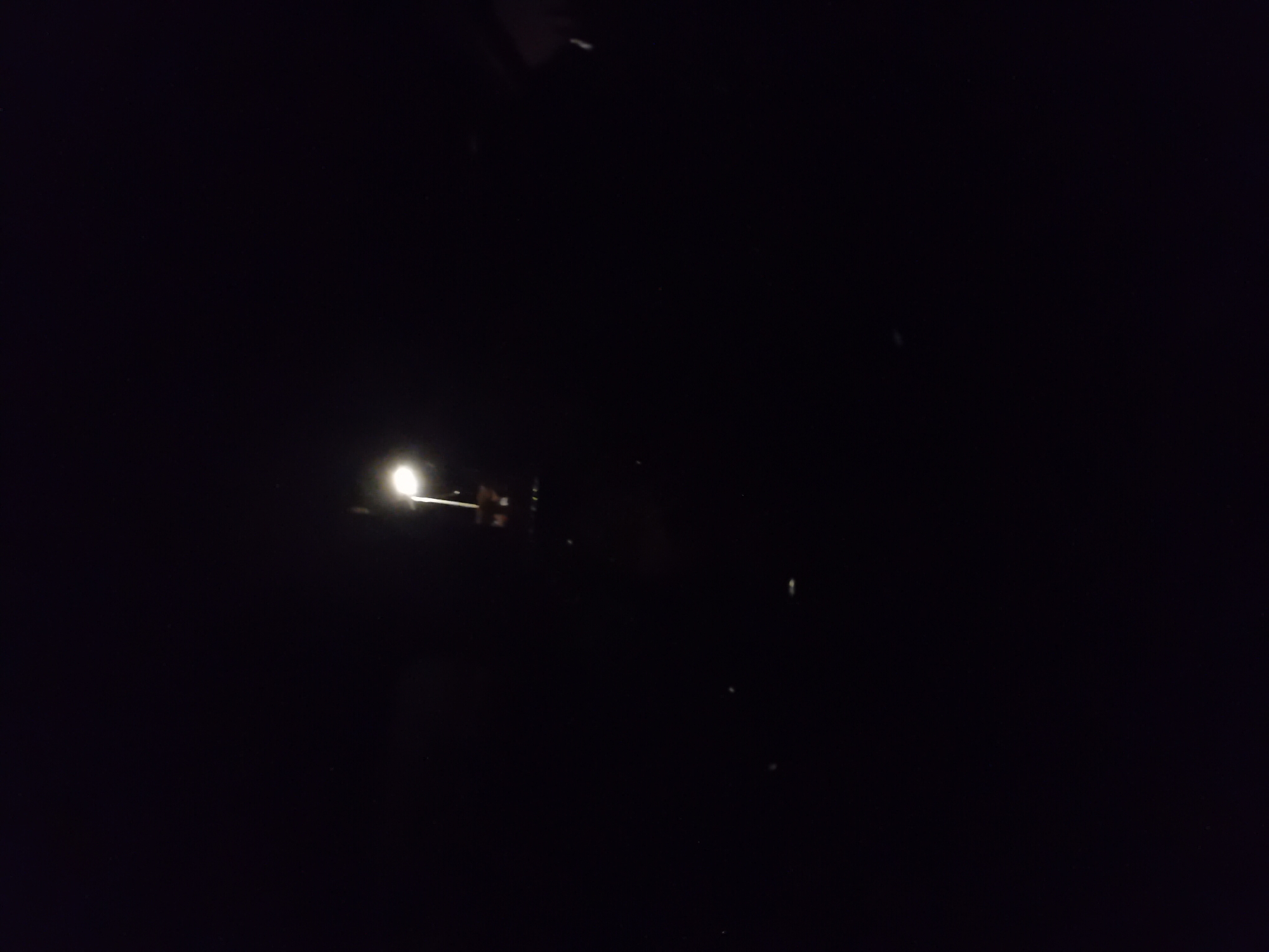 A night-time photograph captures a single streetlamp in the city of Jauro Yinu, Nigeria. The light from the lamp illuminates a small portion of its immediate surroundings, with the rest of the image fading into darkness. The scene appears to be an outdoor urban environment, likely a street or public space, given the presence of the streetlamp. No individuals or specific activities are discernible due to the low light conditions. The overall impression is one of solitude and the stark contrast between the artificial light source and the encompassing dark.